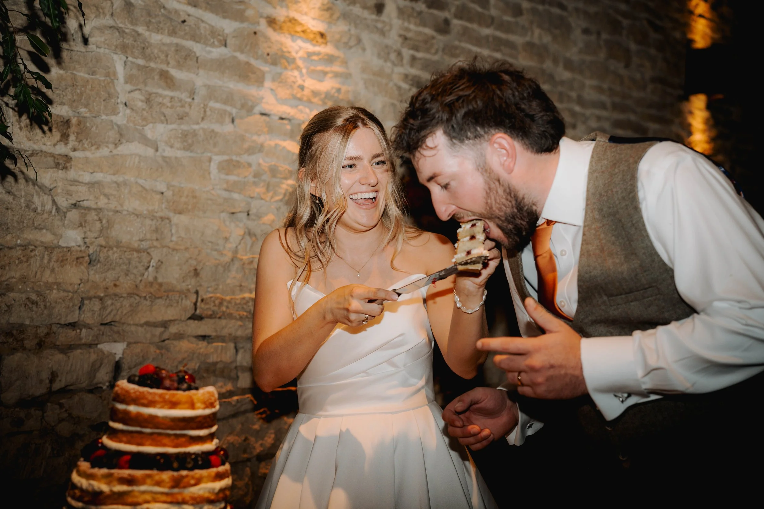 A smiling woman in a white dress happily sharing a slice of birthday cake with a man in a white shirt and waistcoat at a birthday celebration. There is a multi-layered cake on a table in the foreground.