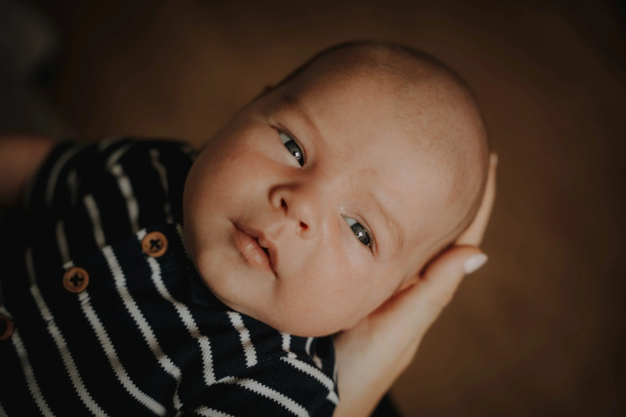 Close-up of a baby with a shaved head lying on a person's hand, wearing a black and white striped shirt with orange buttons.