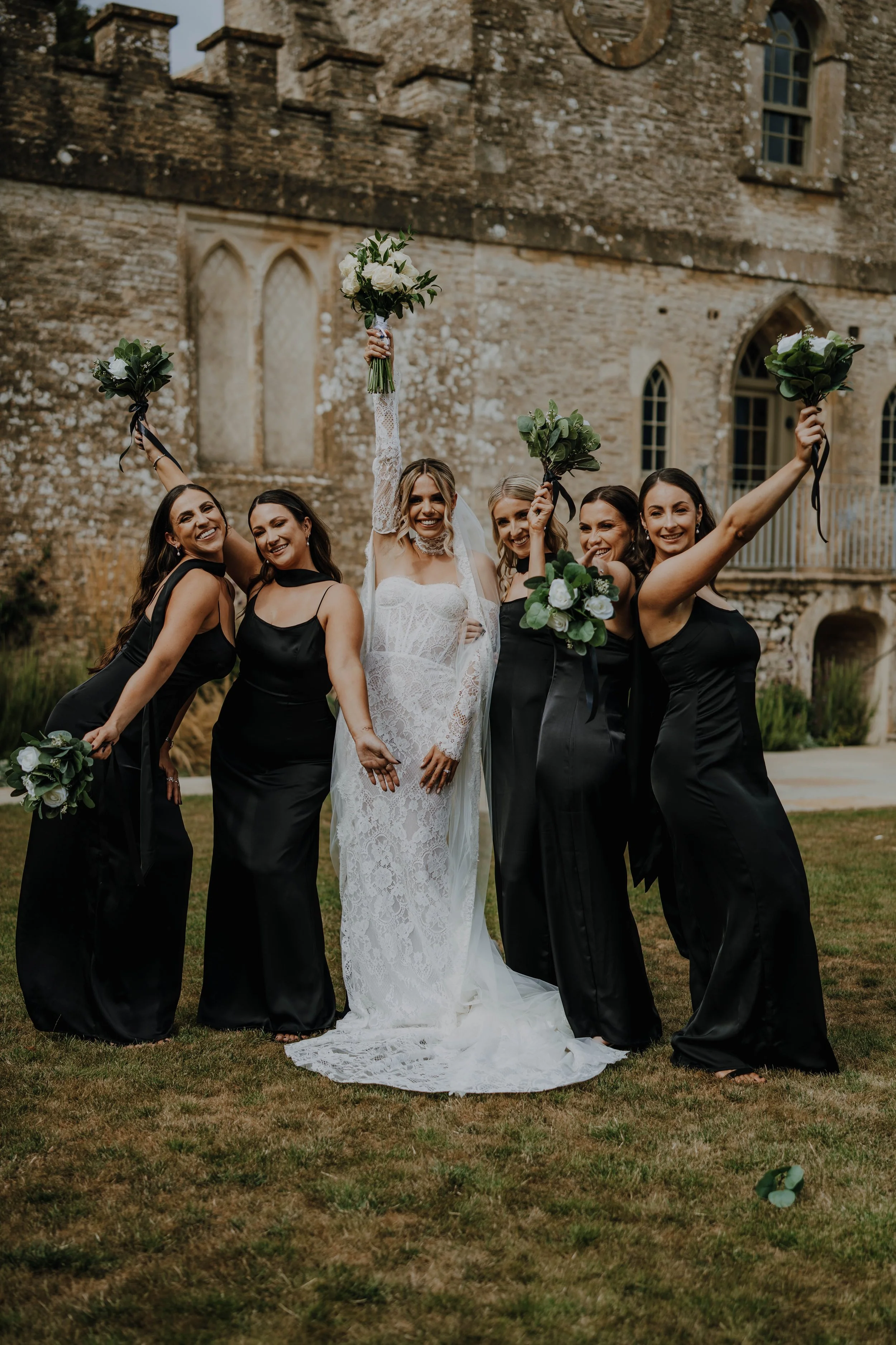 A bride and five bridesmaids celebrating outdoors in front of a historic stone building. The bride is in a white lace wedding dress, holding her bouquet high, with the bridesmaids in black dresses holding their bouquets up in the air.