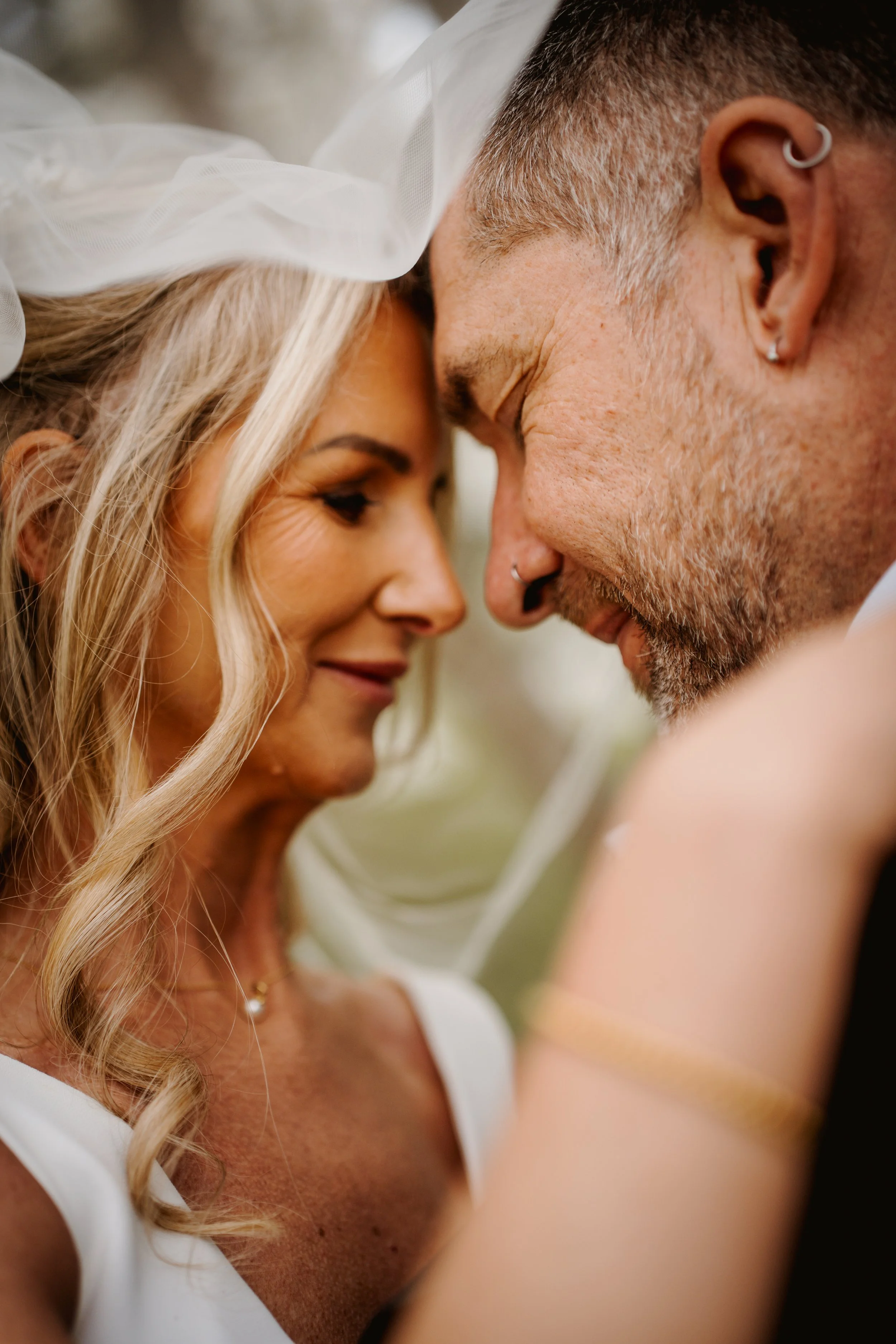 Close-up of a couple with foreheads touching, smiling, with the woman wearing a white veil and the man with a beard and earrings, during a wedding.