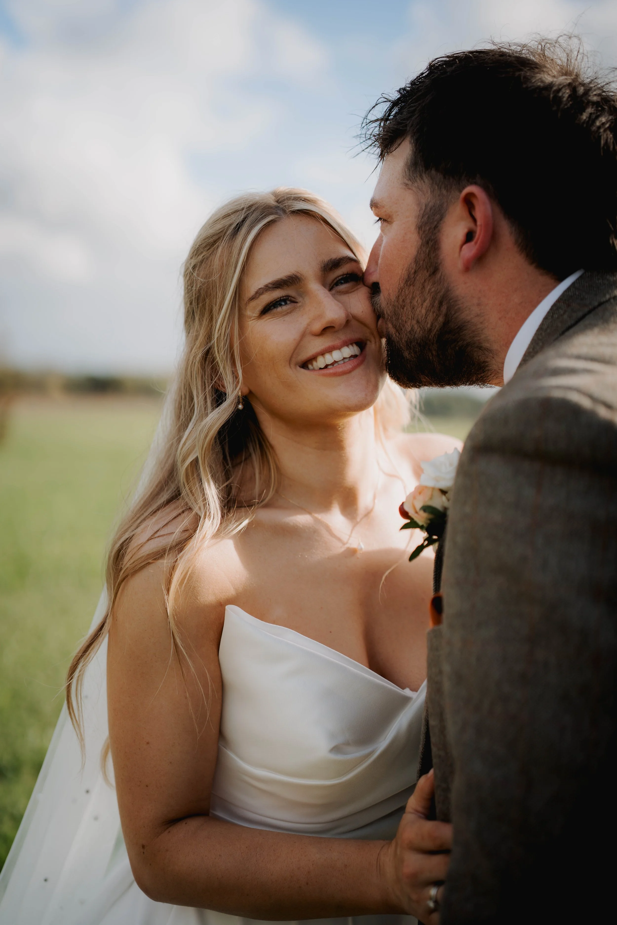 A smiling blonde woman in a white wedding dress looking affectionately at a bearded man in a suit, who is kissing her cheek outdoors under a cloudy sky.