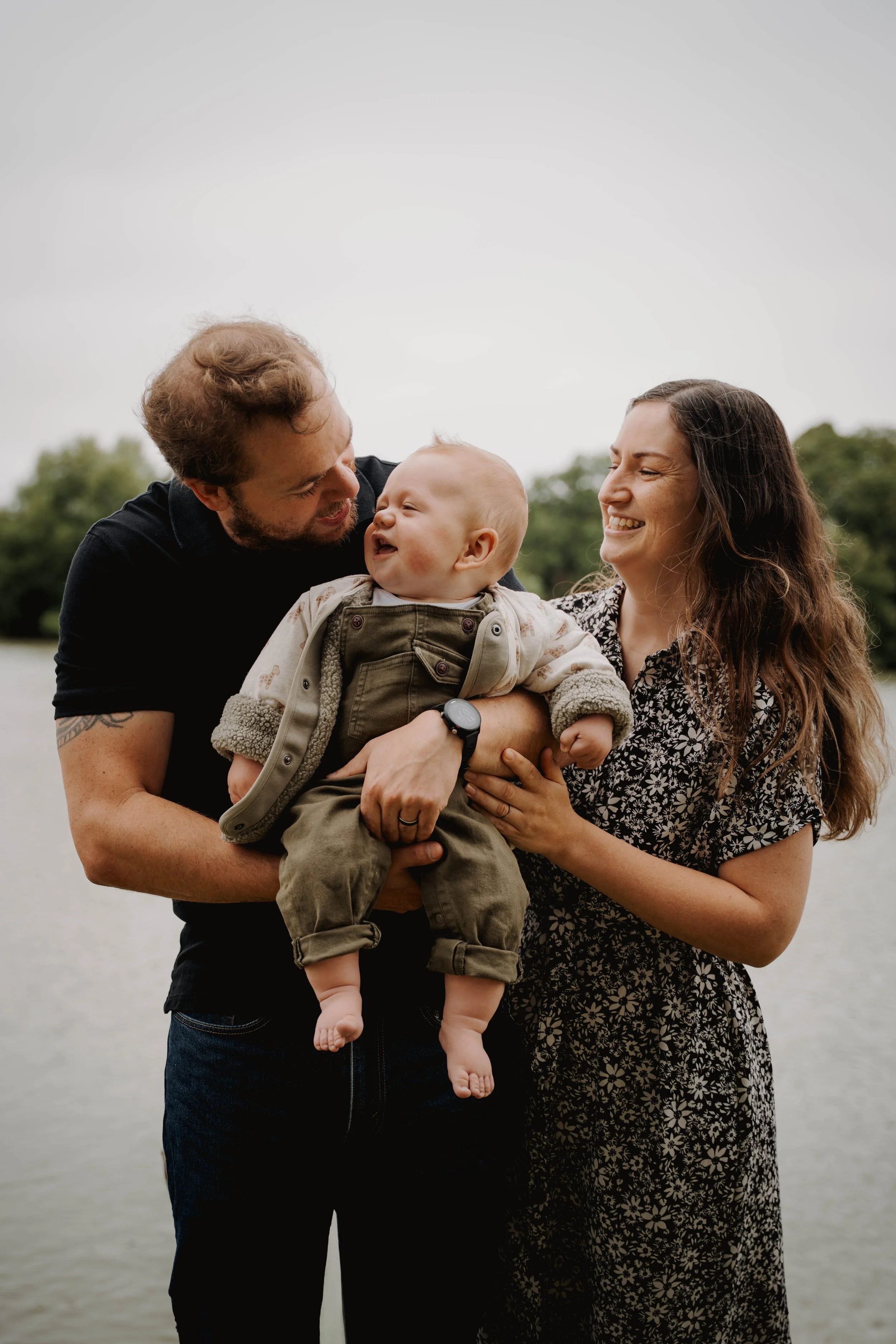 A happy family of three: a man, woman, and a baby, outdoors near a body of water with trees in the background, smiling and enjoying the moment.
