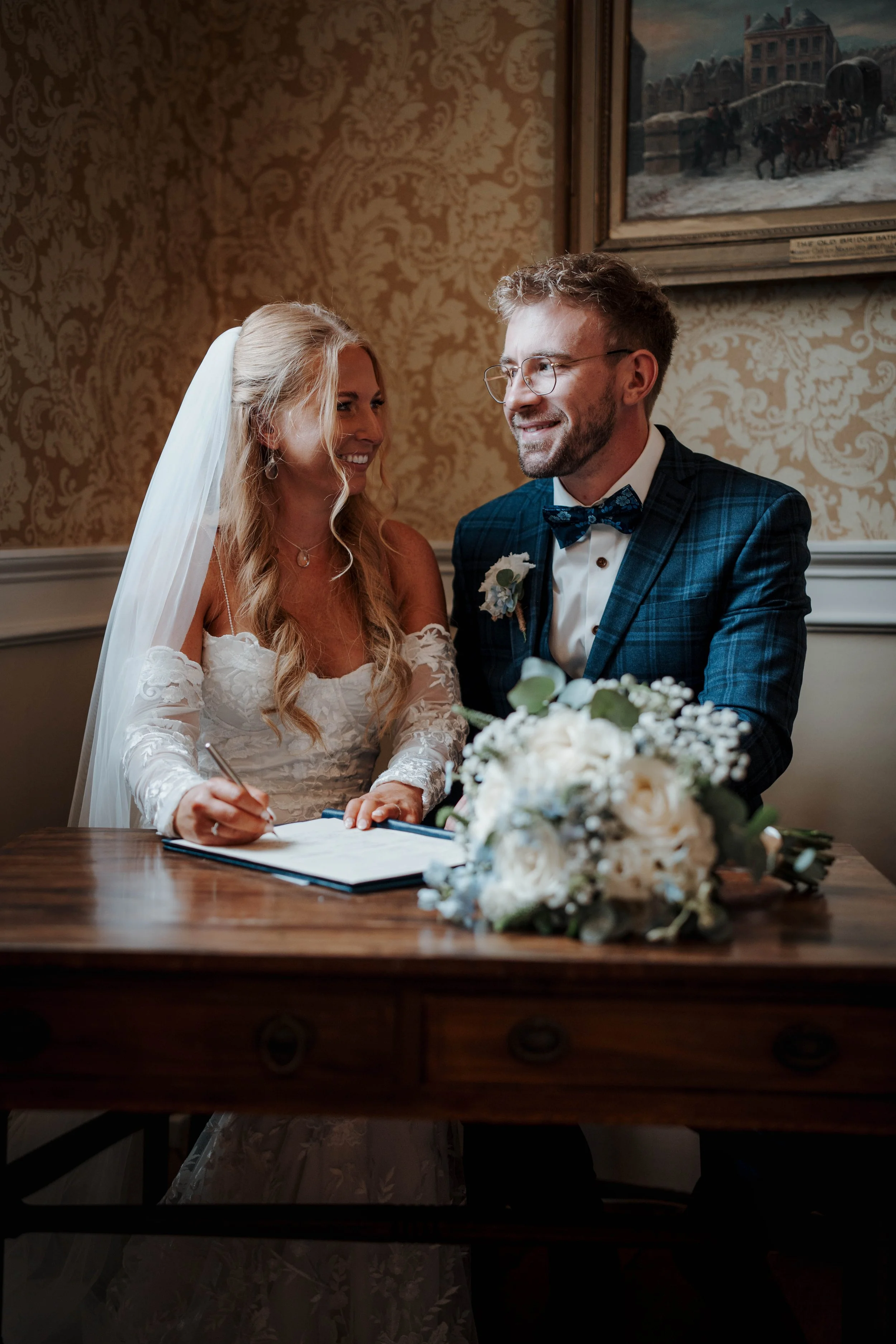 A bride and groom signing a marriage certificate during their wedding ceremony, sitting at a wooden table with a floral arrangement in front of them, in a decorated indoor setting.
