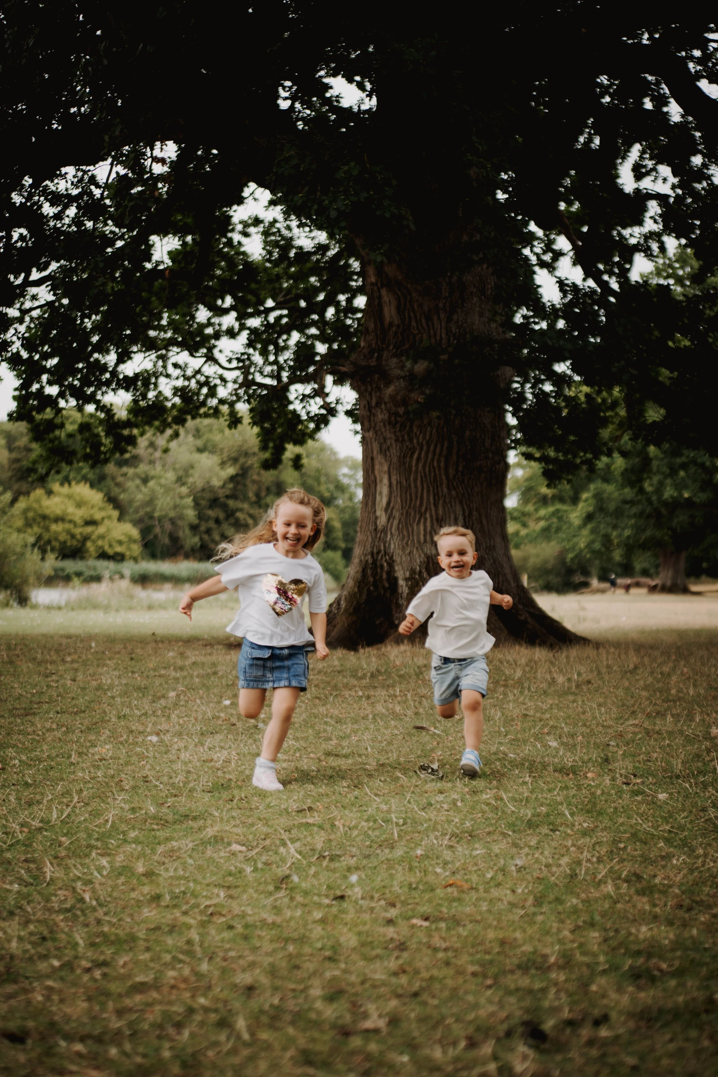 Two children, a girl and a boy, running and playing outdoors near a large tree.