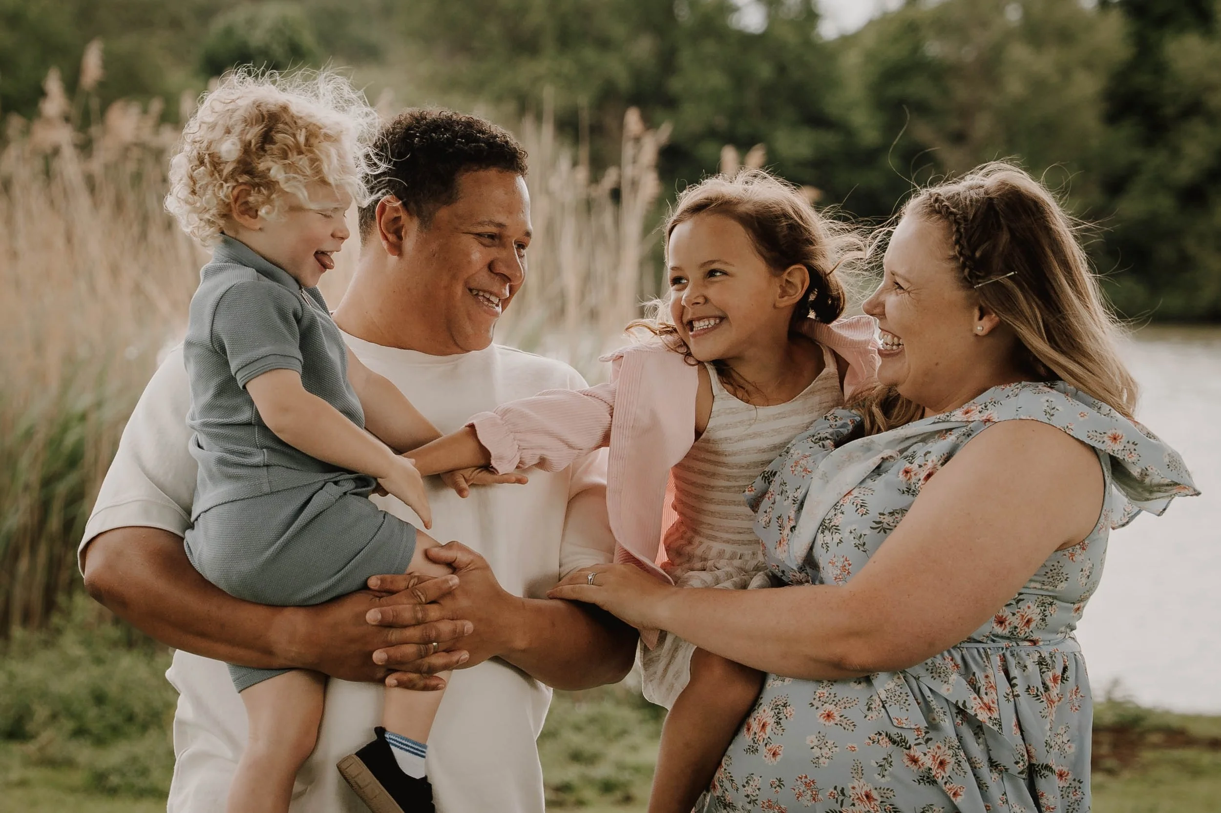A diverse family of five enjoying time outdoors near a body of water, smiling and touching each other.