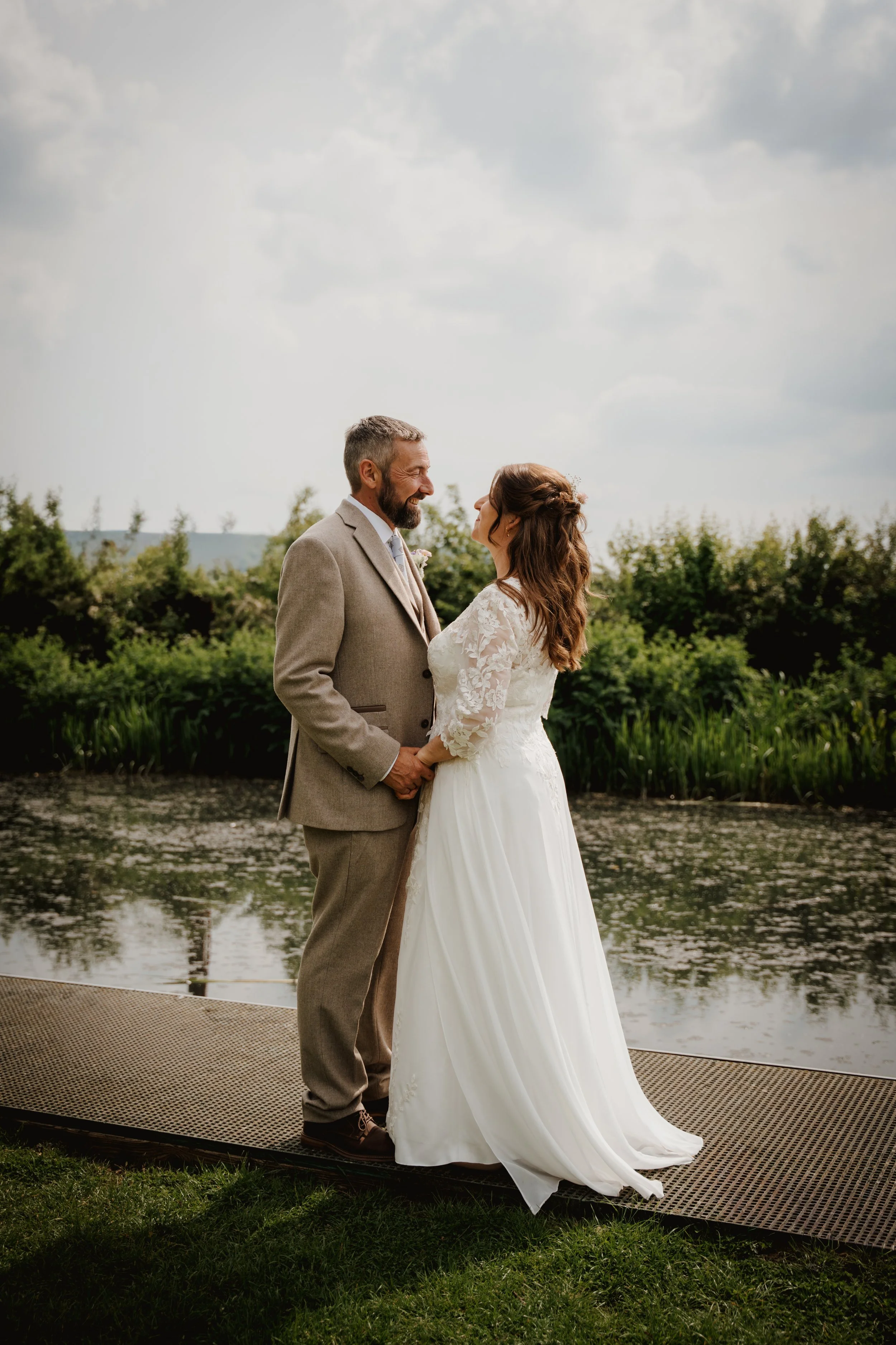 A bride and groom standing hand in hand outdoors, gazing at each other, with a pond and green trees in the background on a cloudy day.