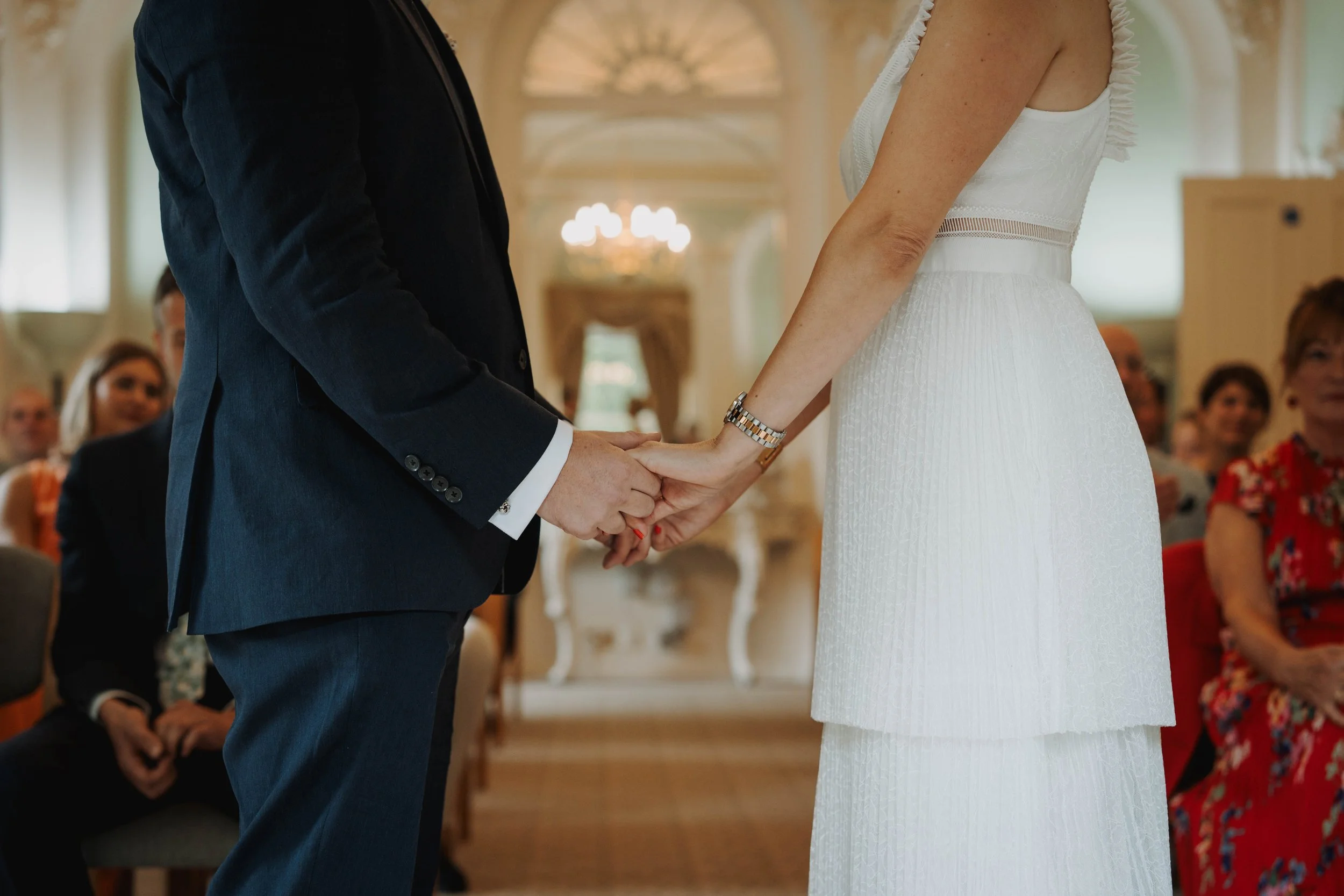 A close-up of a couple holding hands during a wedding ceremony, with the bride in a white dress and the groom in a dark suit, inside a decorated venue with seated guests watching.