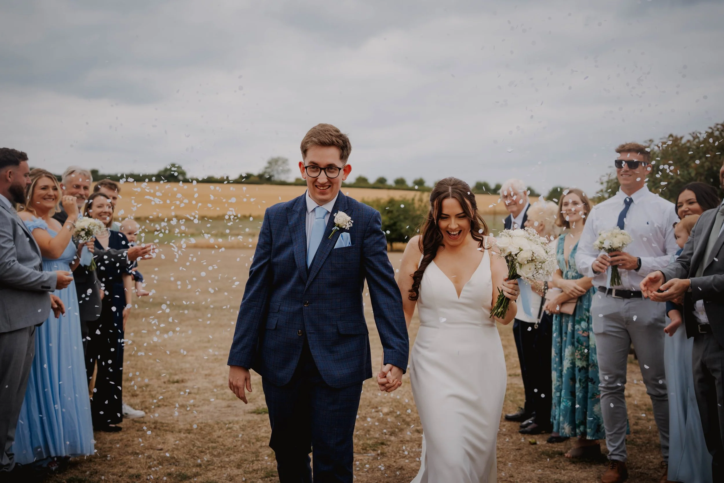 A newlywed couple holding hands and walking through a confetti celebration outdoors with friends and family on a cloudy day.