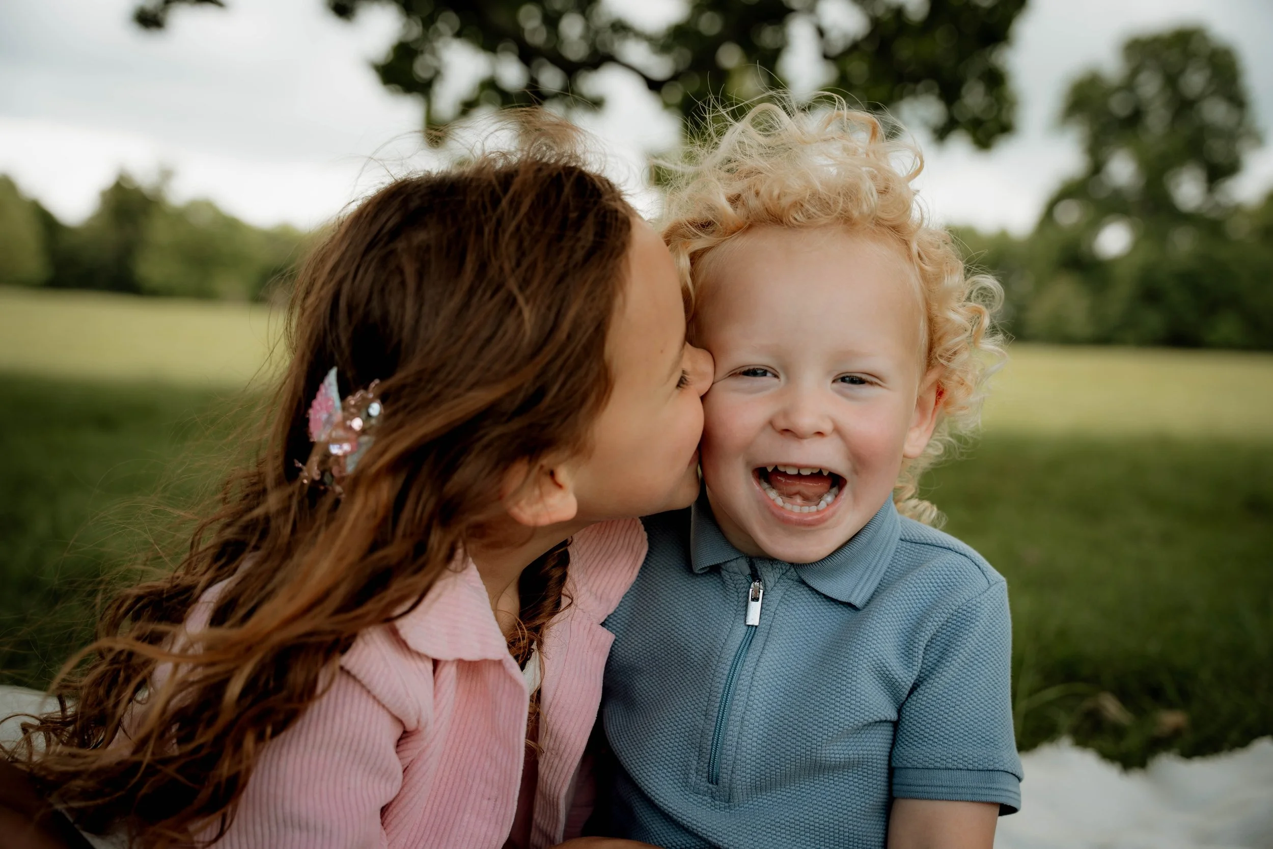 A woman with long red hair leaning in to kiss a young boy on the cheek, both are smiling outdoors with a blurred background of trees and grass.