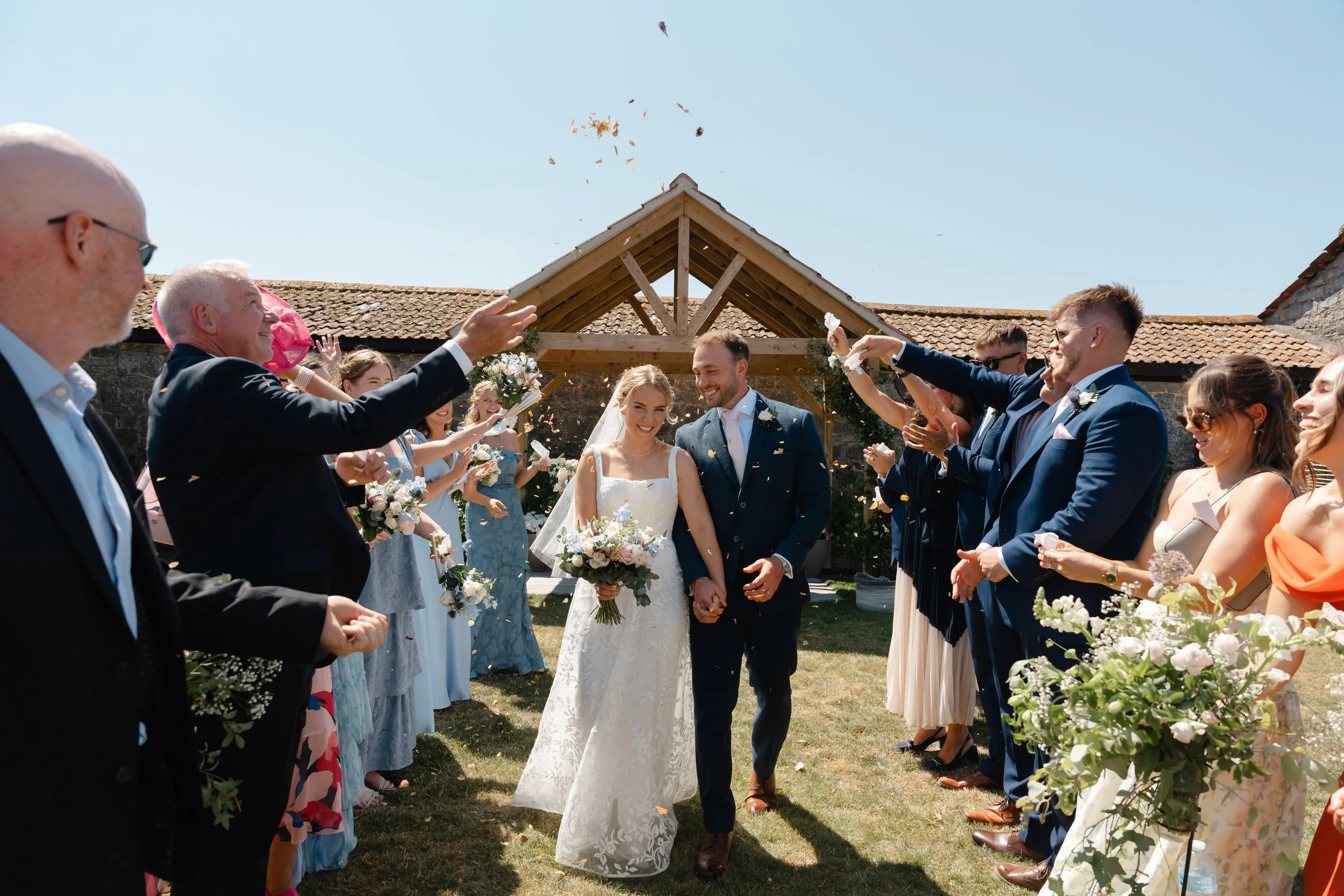 A bride and groom smiling and holding hands during their outdoor wedding ceremony, surrounded by guests throwing flower petals.