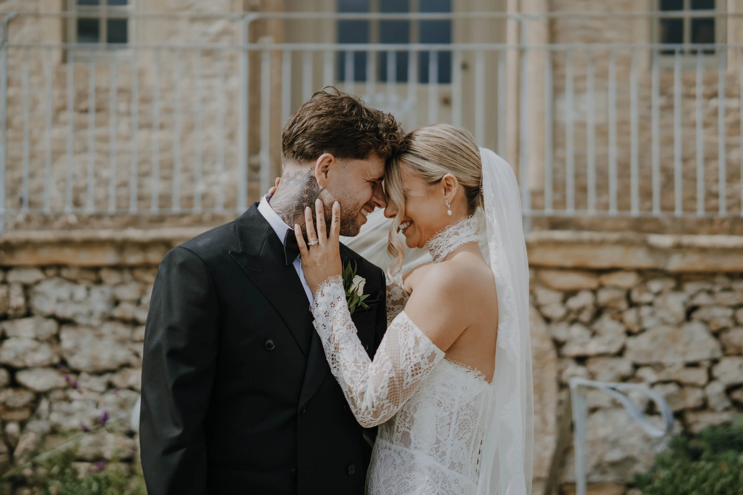 A bride and groom are smiling and touching foreheads outdoors with a stone wall and metal fence in the background.