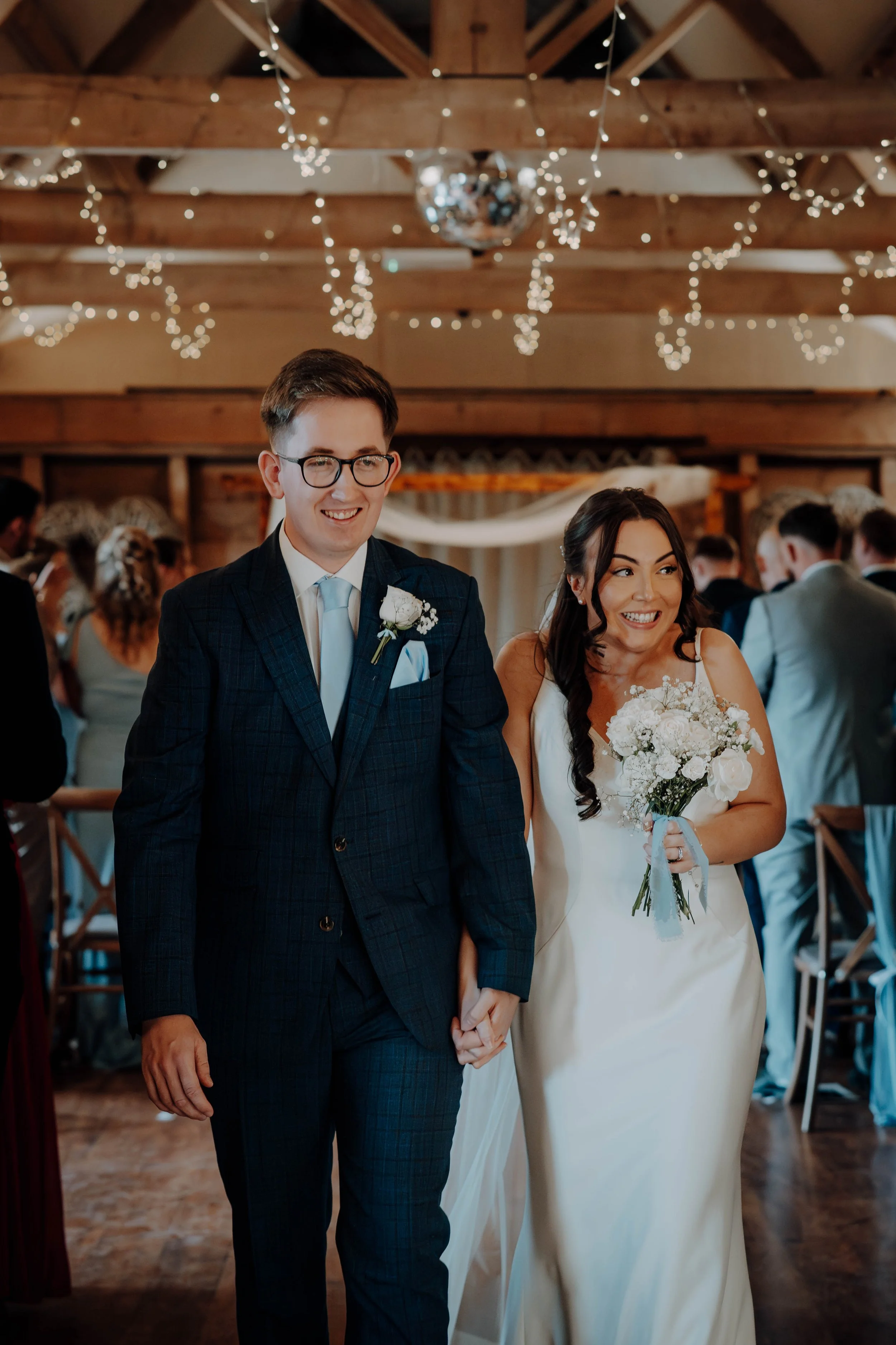 A bride and groom walking hand in hand at their wedding reception, smiling. The groom wears glasses and a navy blue suit with a light blue tie, and the bride wears a white wedding dress and holds a bouquet of white flowers. The reception occurs in a 