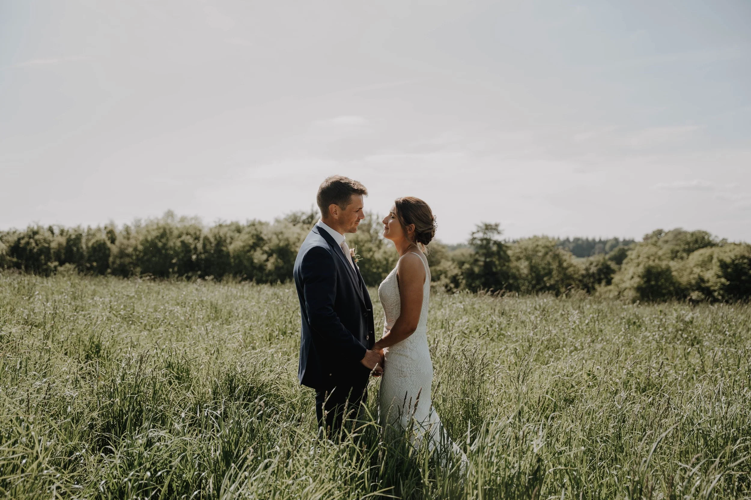 A bride and groom stand in a grassy field, facing each other and holding hands, with a background of trees and a partly cloudy sky.