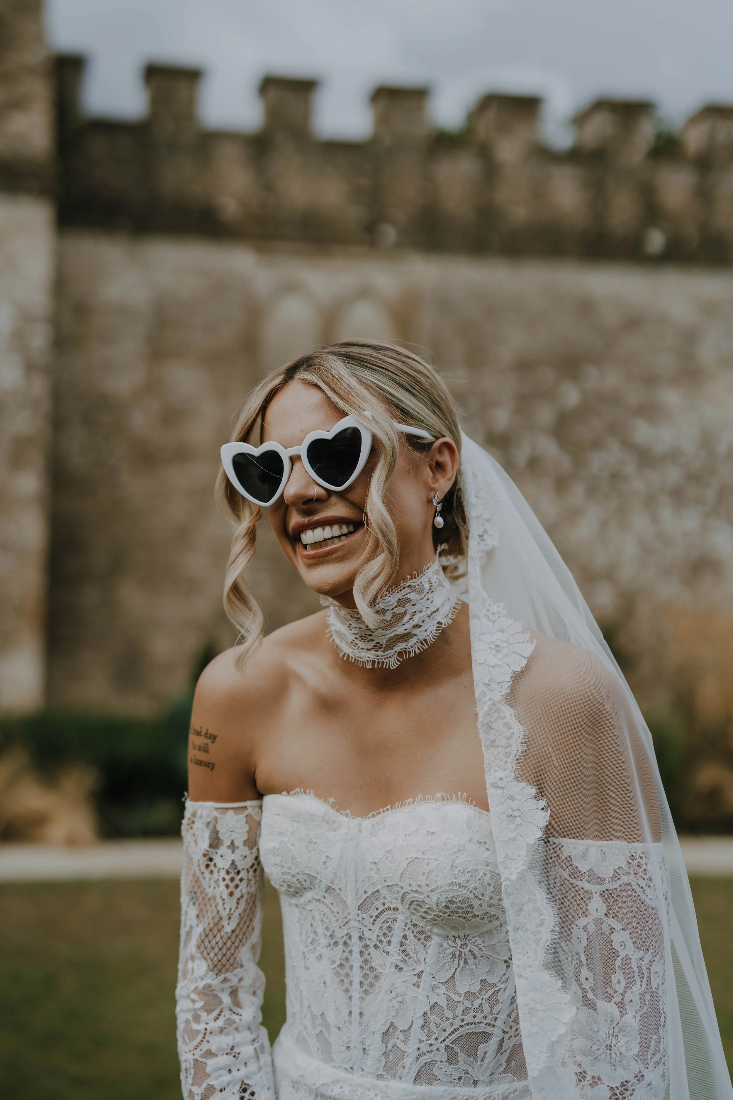 Bride wearing heart-shaped sunglasses, lace wedding dress, and a lace choker, laughing outdoors with a castle wall in the background.