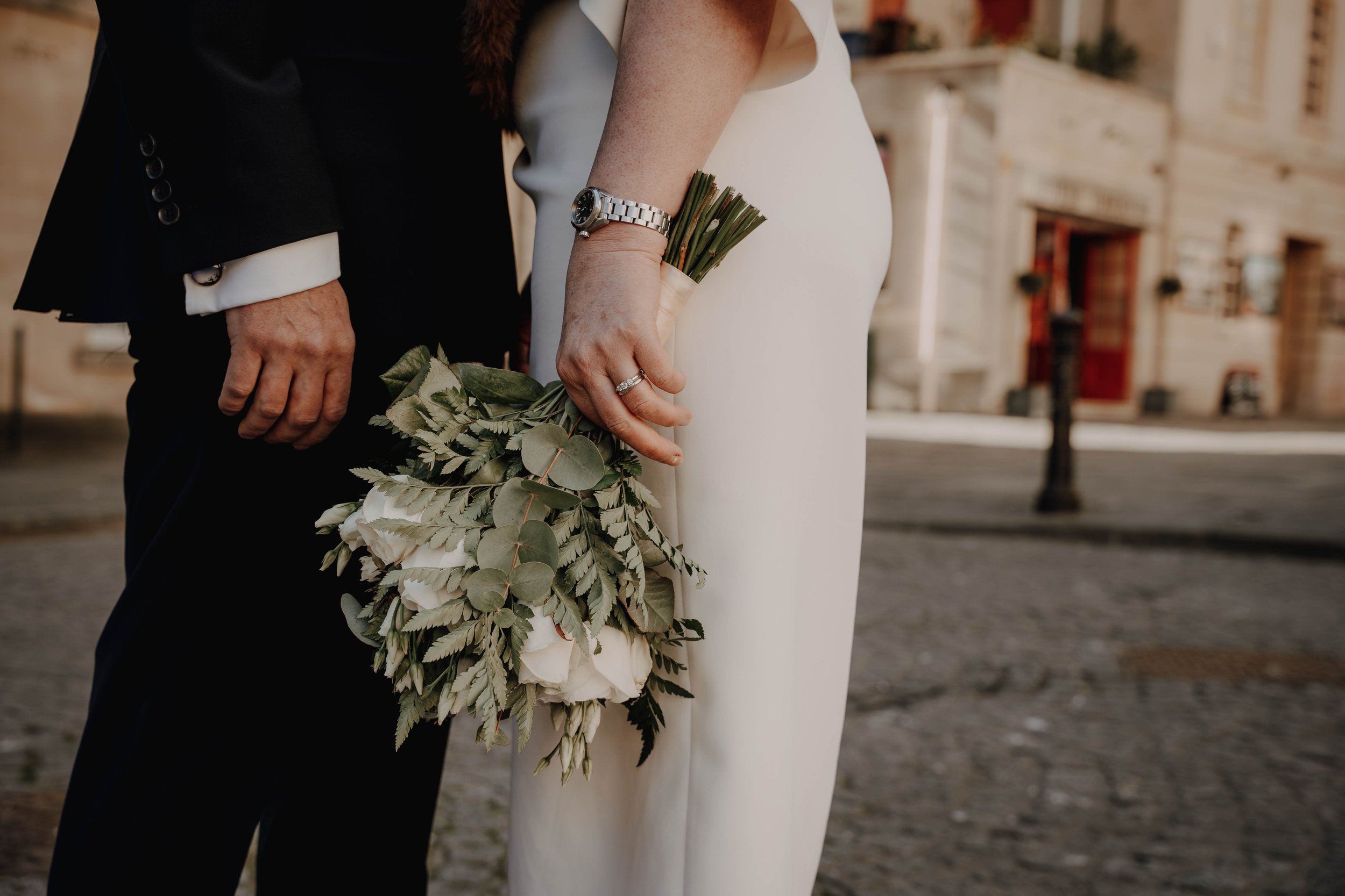 Close-up of a couple holding hands, with one holding a bouquet of greenery, in an urban setting with buildings in the blurred background.