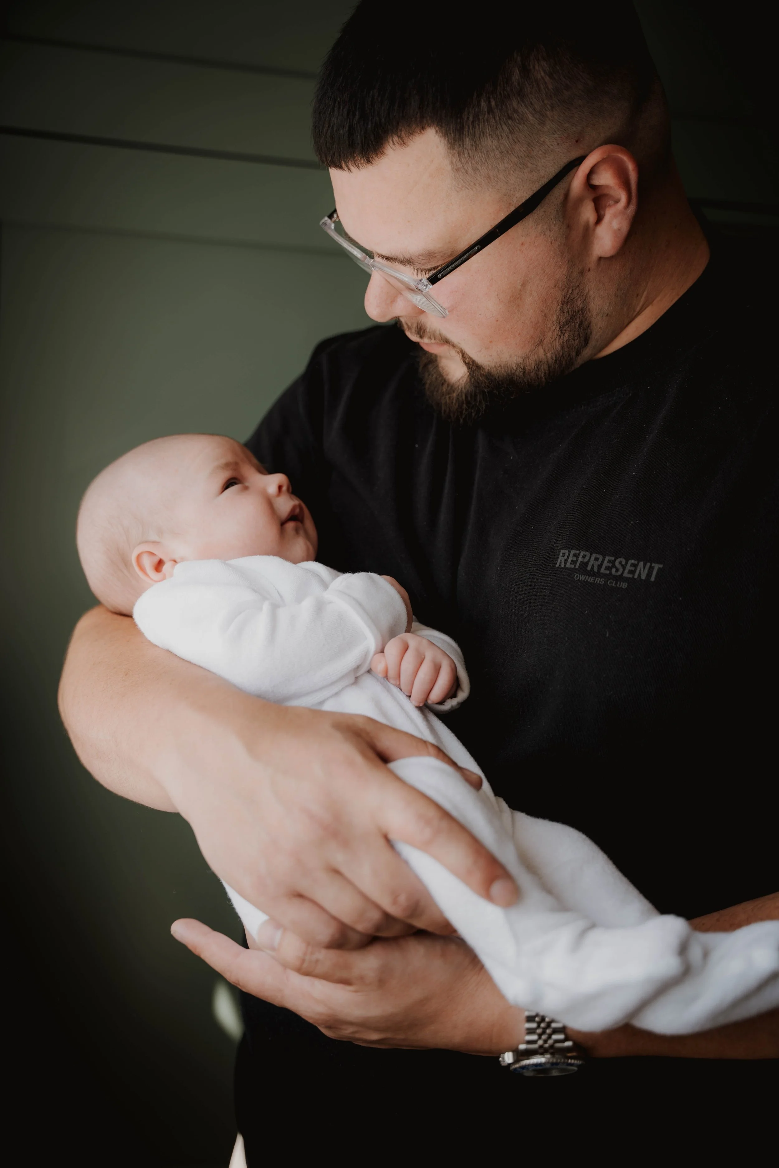 A man with glasses and a beard holding a baby in a white outfit, looking at each other lovingly indoors.