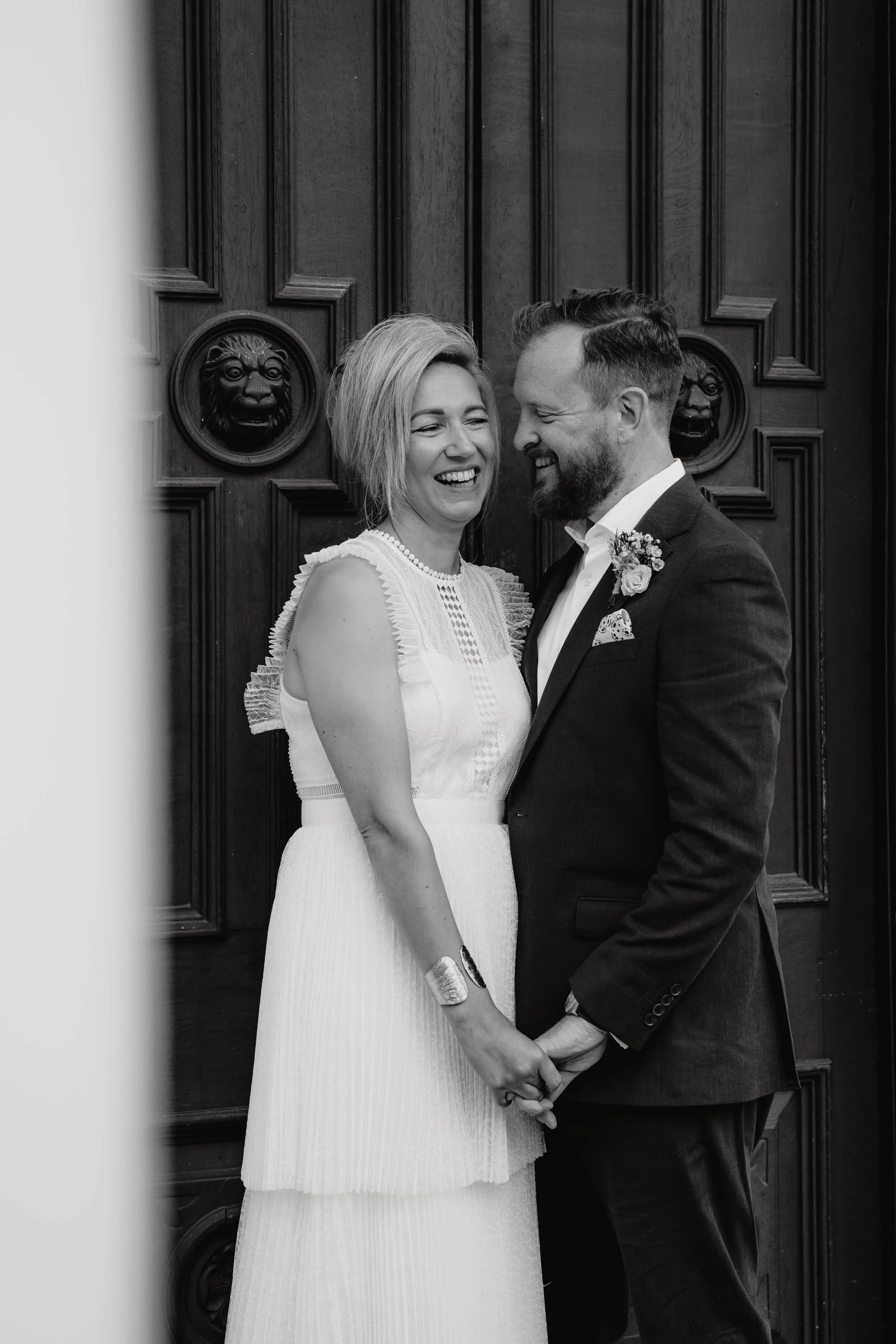 A black and white photo of a smiling couple dressed in wedding attire, standing close together and holding hands in front of a dark wooden door with lion face carvings.