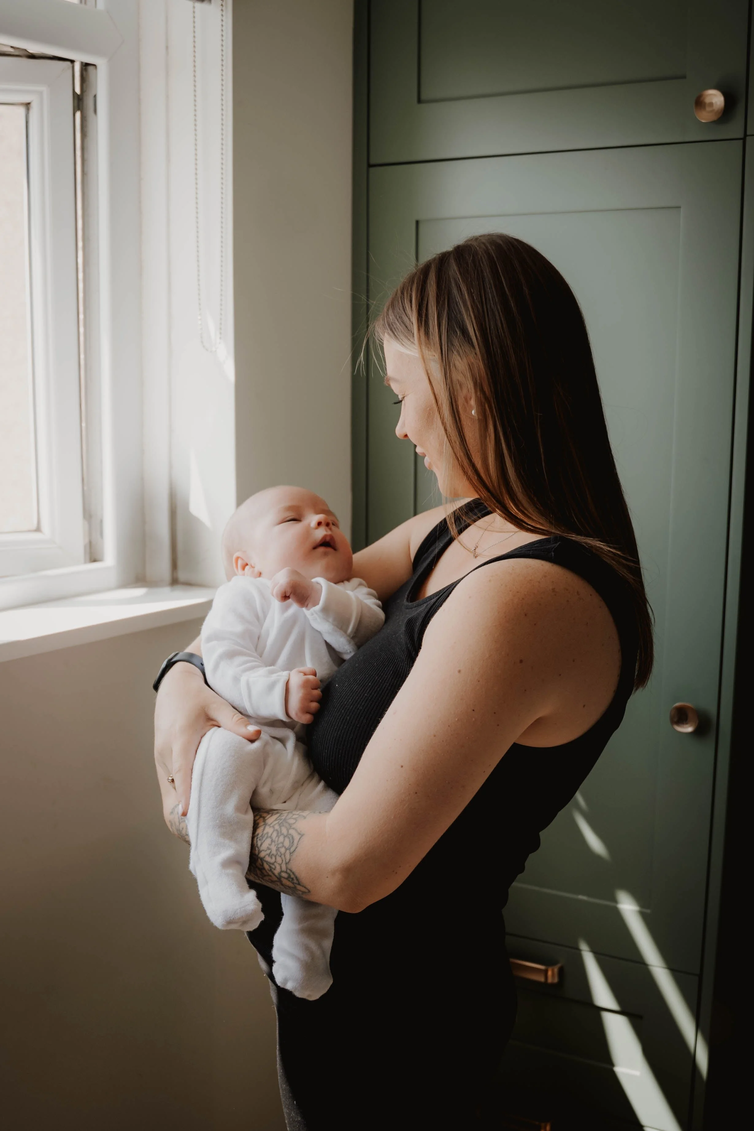 A woman holding a baby near a window in a kitchen with green cabinets.
