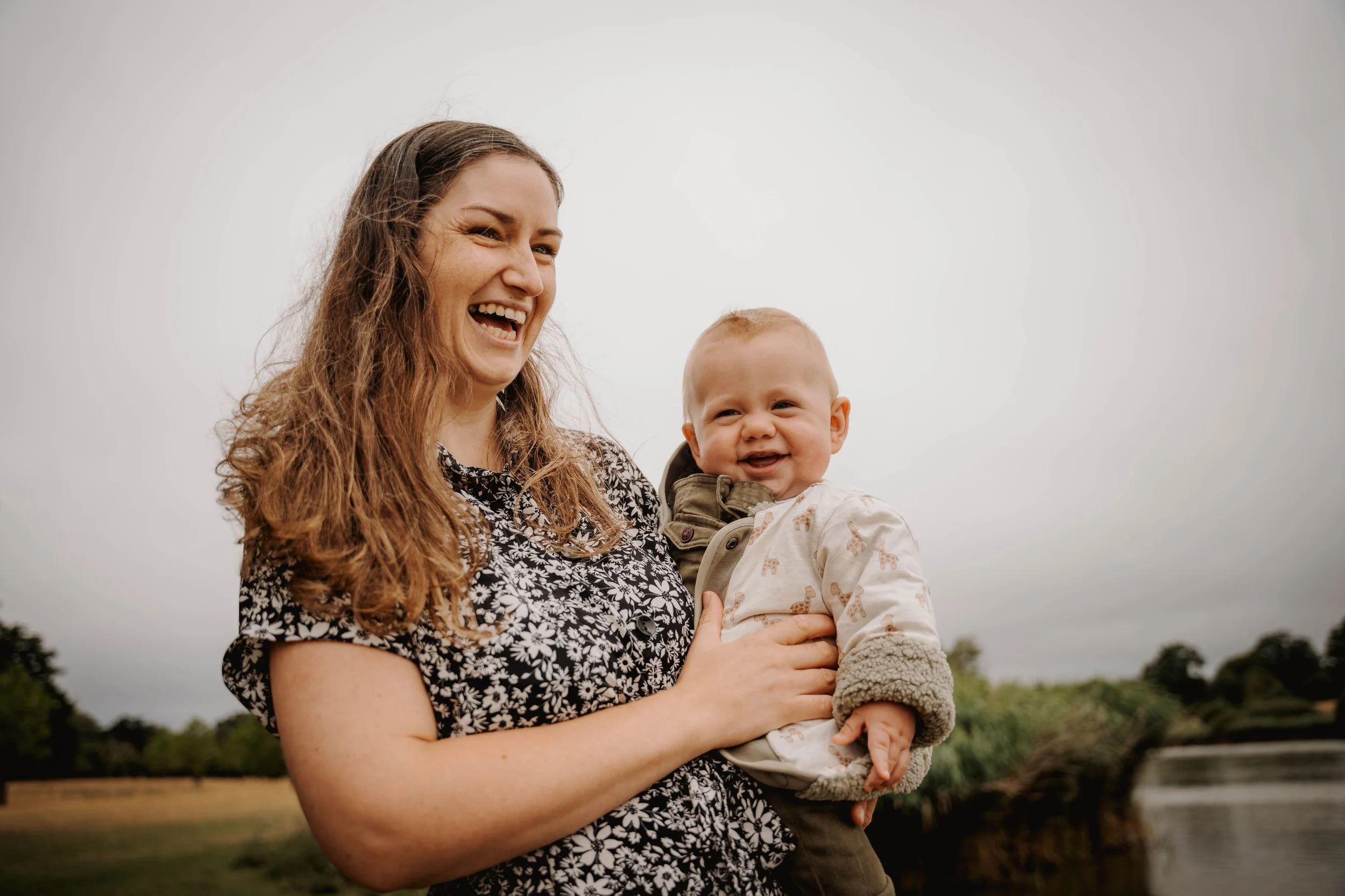 A woman with long, wavy brown hair holding a smiling baby outside near a water body and trees on a cloudy day.