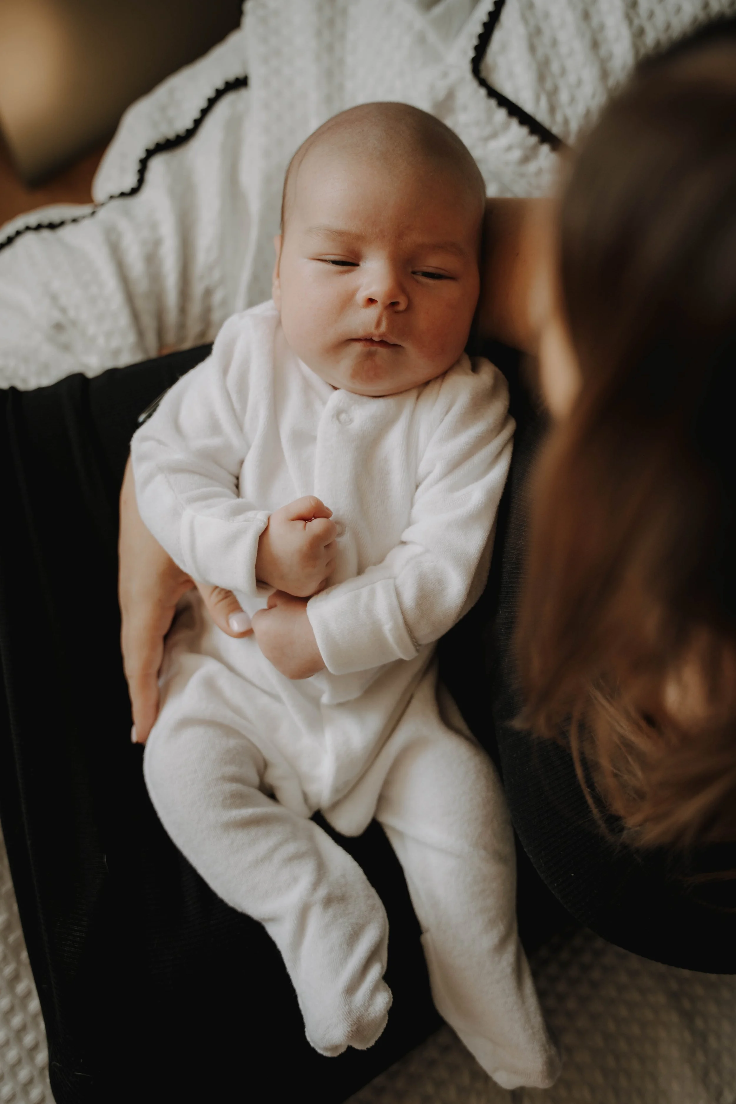 Close-up of a baby with a round face and closed eyes, wearing a white onesie, sitting on an adult's lap.