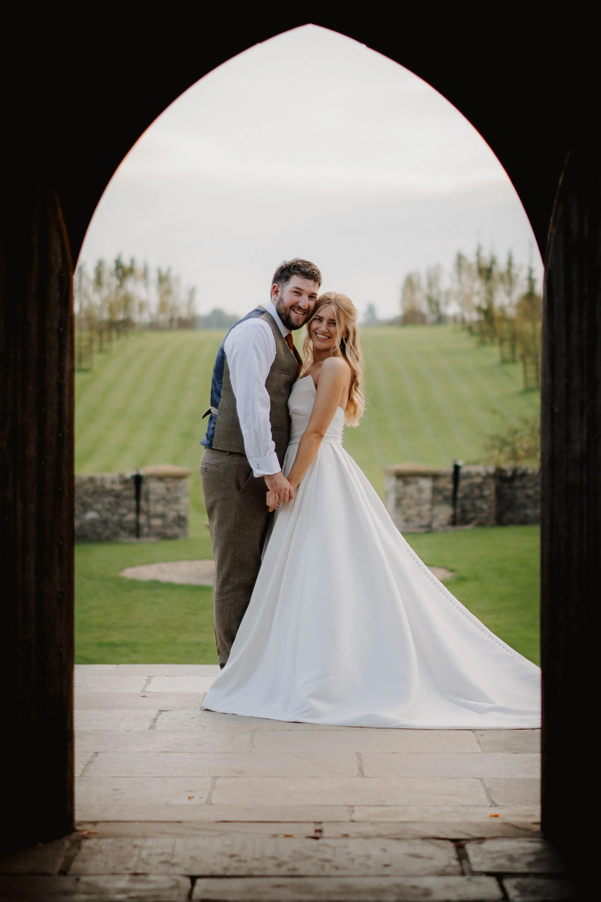 A newlywed couple standing under an archway at a park, holding hands and smiling, with the bride in a white wedding gown and the groom in a gray suit and tie, with trees and a grassy field in the background.