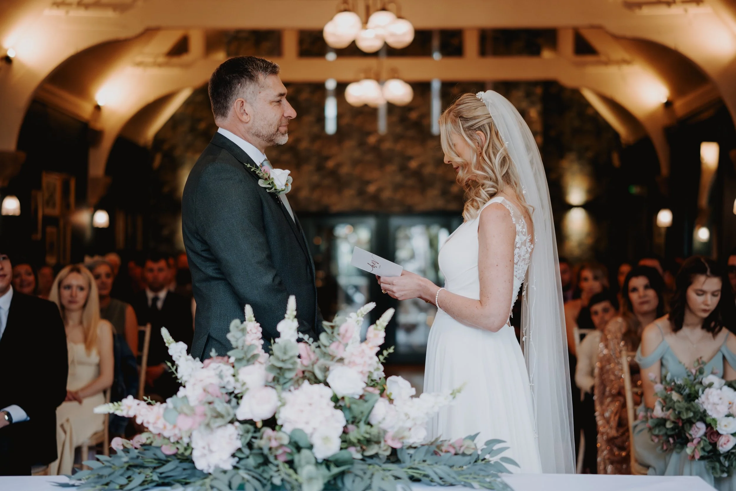 A bride and groom exchange vows during a wedding ceremony inside a warmly lit venue. The bride has long blonde curly hair and wears a white wedding dress with lace details and a veil. The groom has short hair and a beard, wearing a dark suit with a b