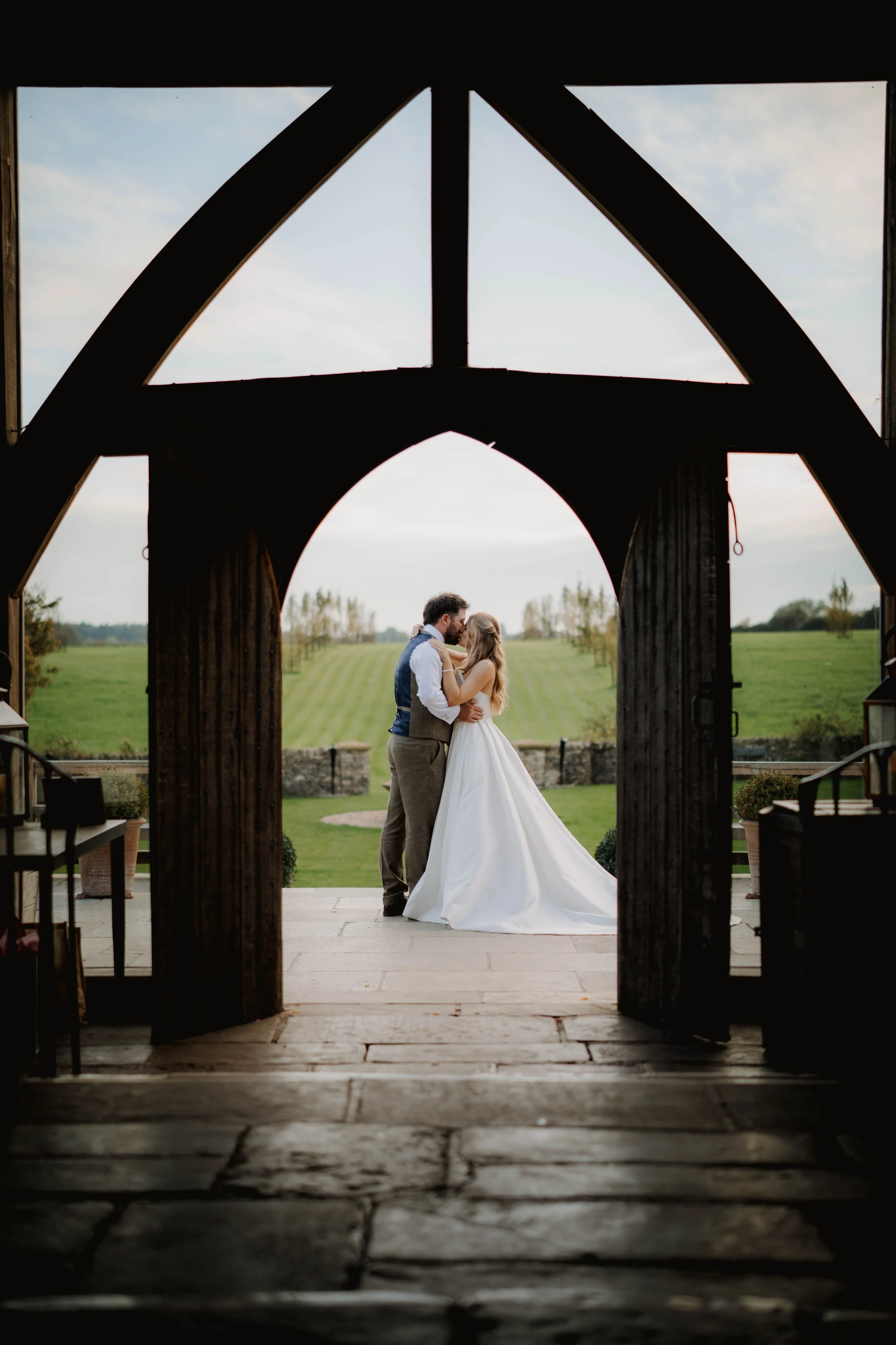 A couple in wedding attire embracing beneath a rustic wooden archway, with green fields and trees in the background.