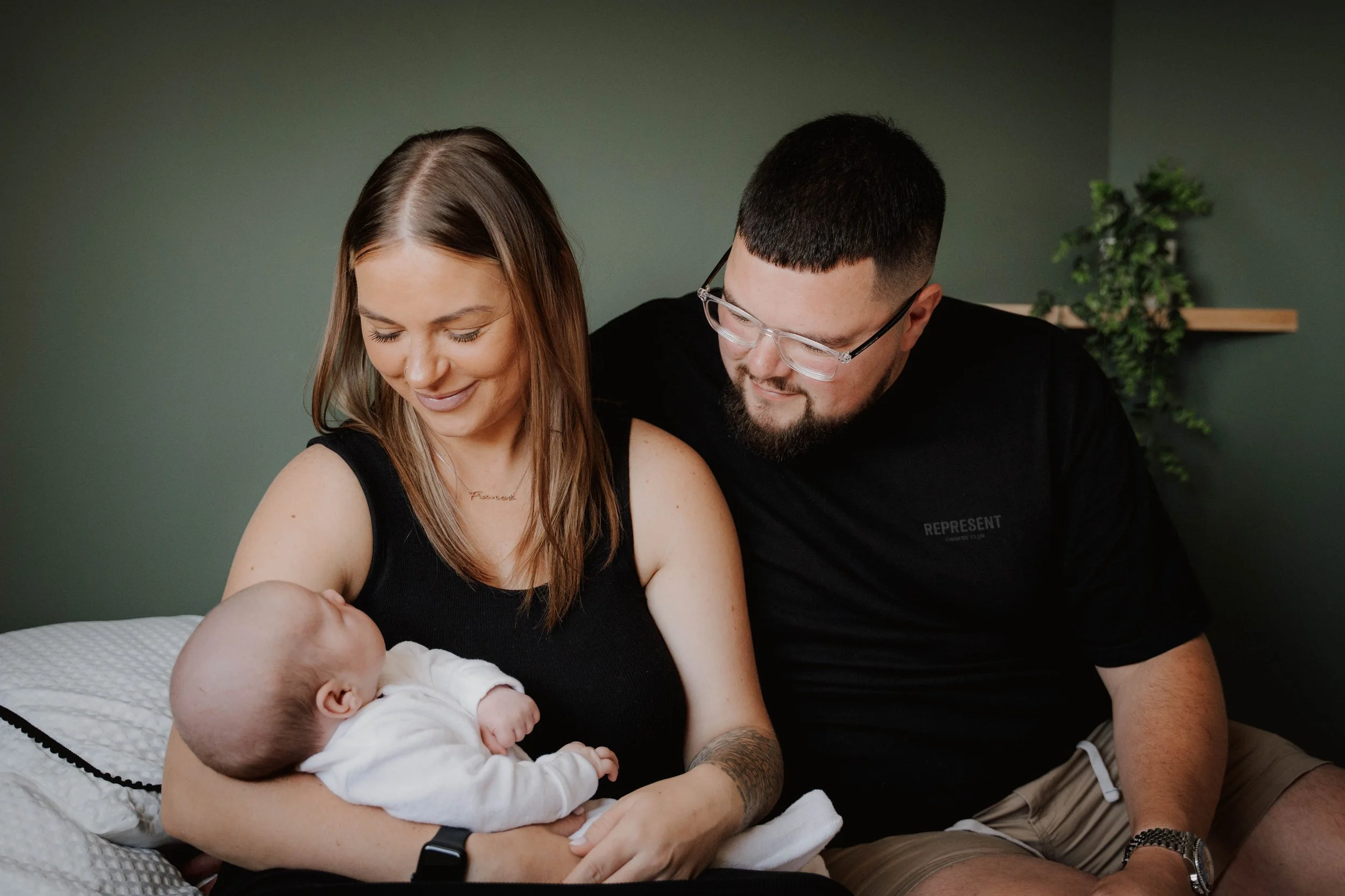 A woman holding a sleeping baby, with a man sitting beside her, looking at the baby with a smile, in a cozy indoor setting.