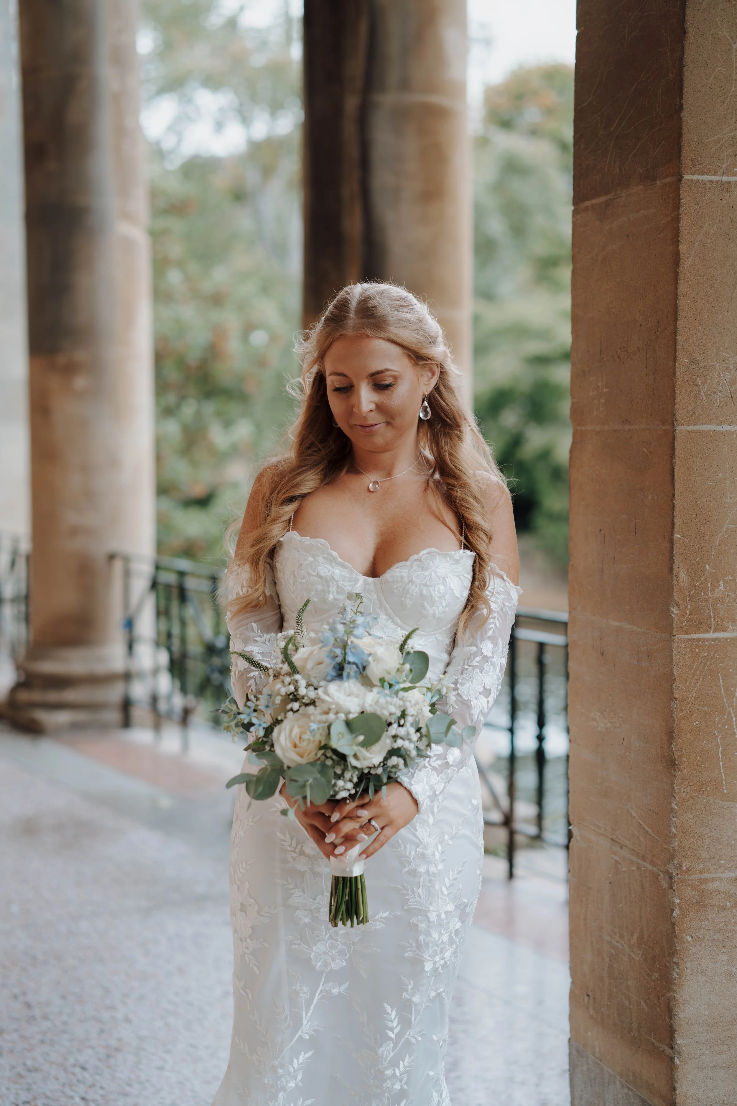 A woman in a white lace wedding dress holding a bouquet of white and light blue flowers, standing outdoors near a stone pillar with blurred trees in the background.