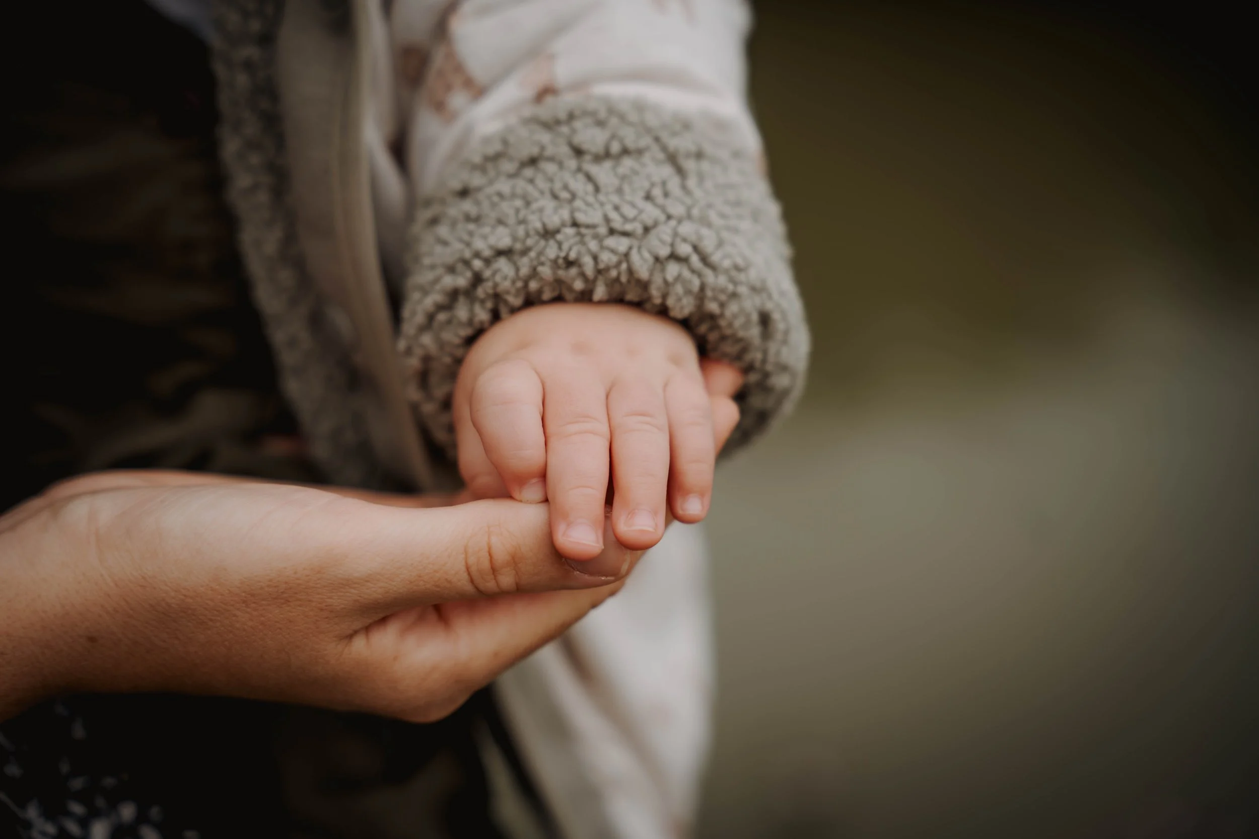 Close-up of a small child's hand holding an adult's finger, dressed in a cozy grey sleeve and outdoors with a blurred background.