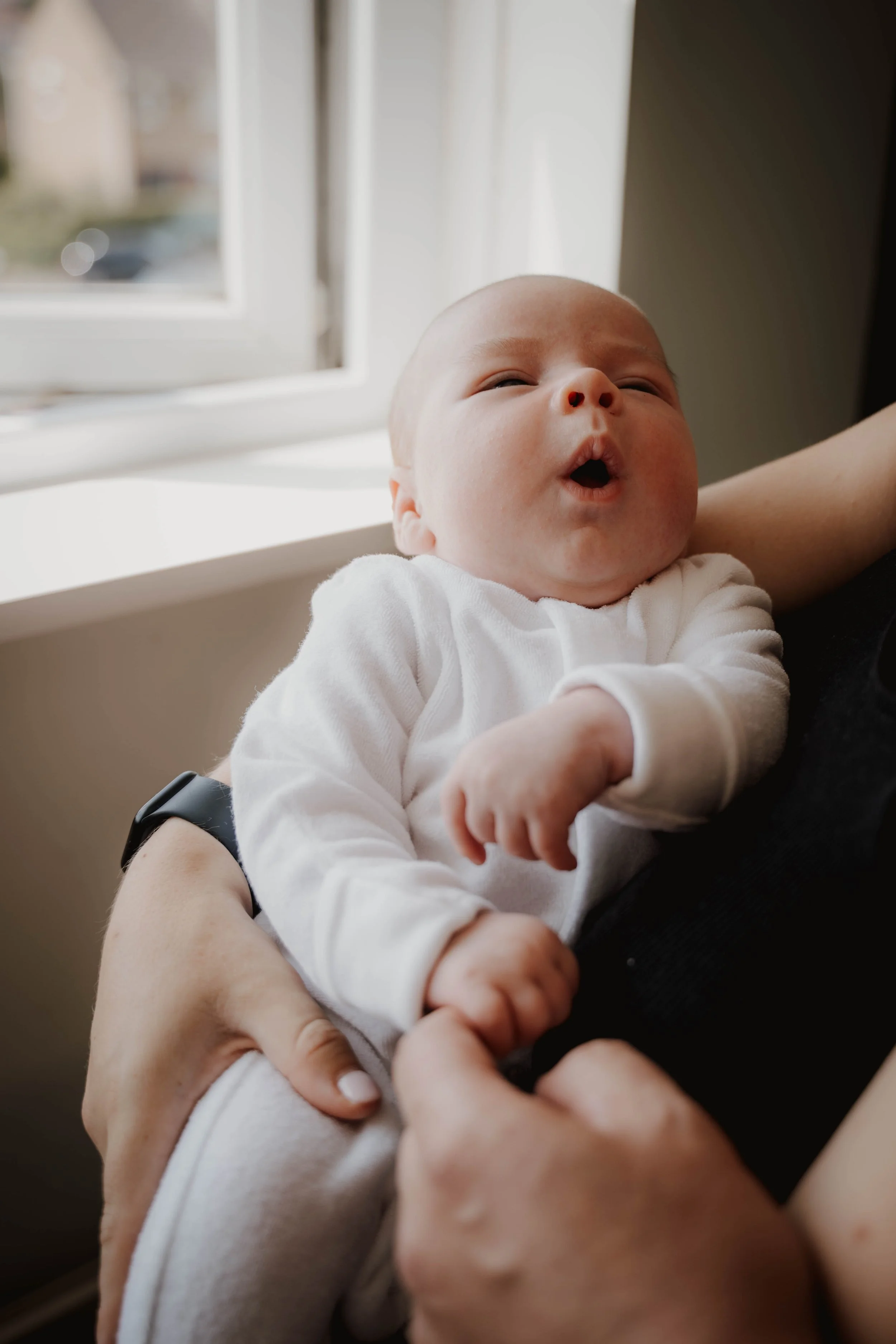 A baby with a light complexion and a bald head, dressed in a white onesie, being held near a window with a view of a blurred outdoor scene, making an funny facial expression with lips puckered.