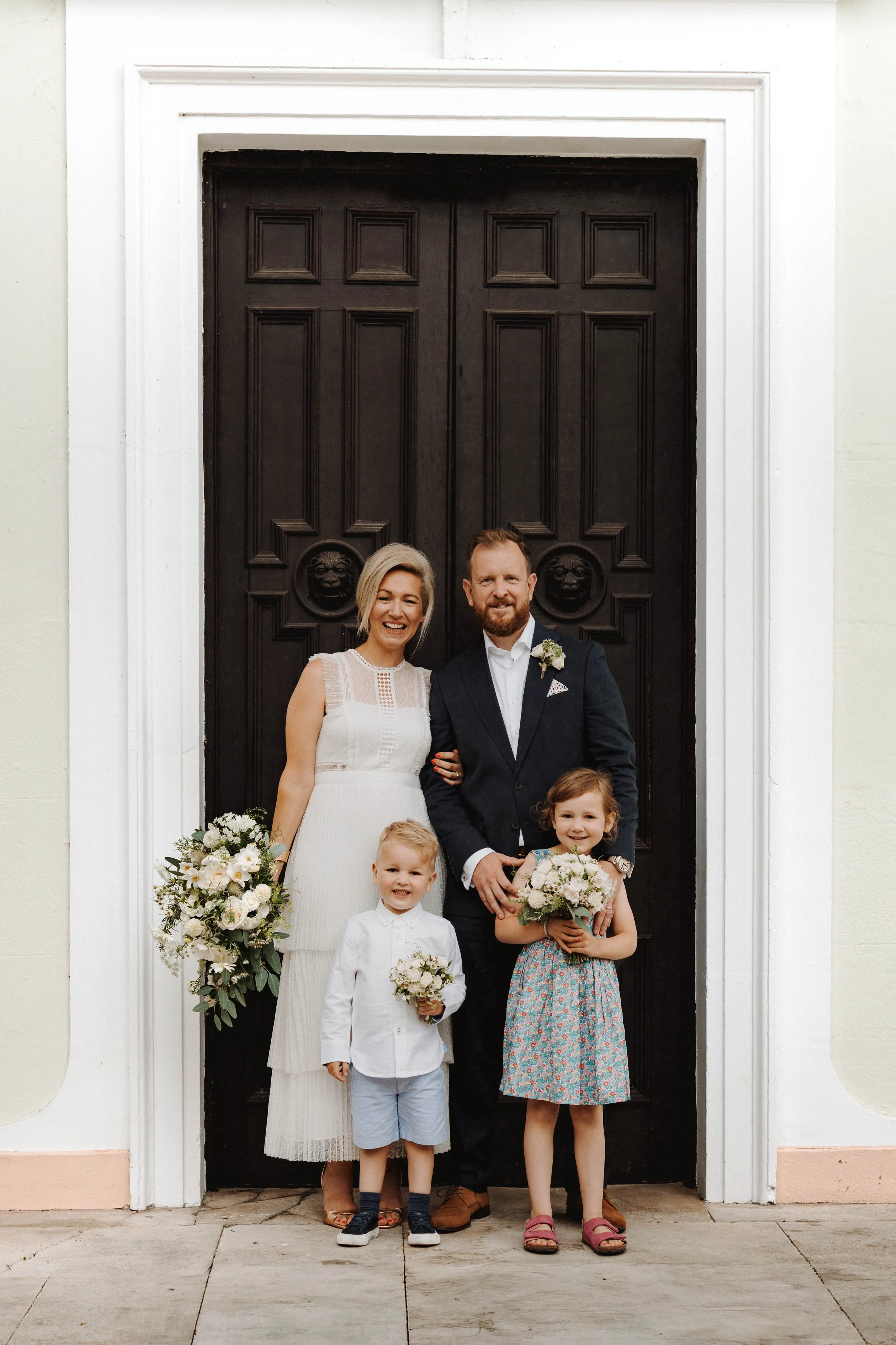 A family of six, including a woman in a white dress, a man in a dark suit, and four children, standing in front of a large dark wooden door with lion head knockers, outside a white building. They are smiling and holding flower bouquets.