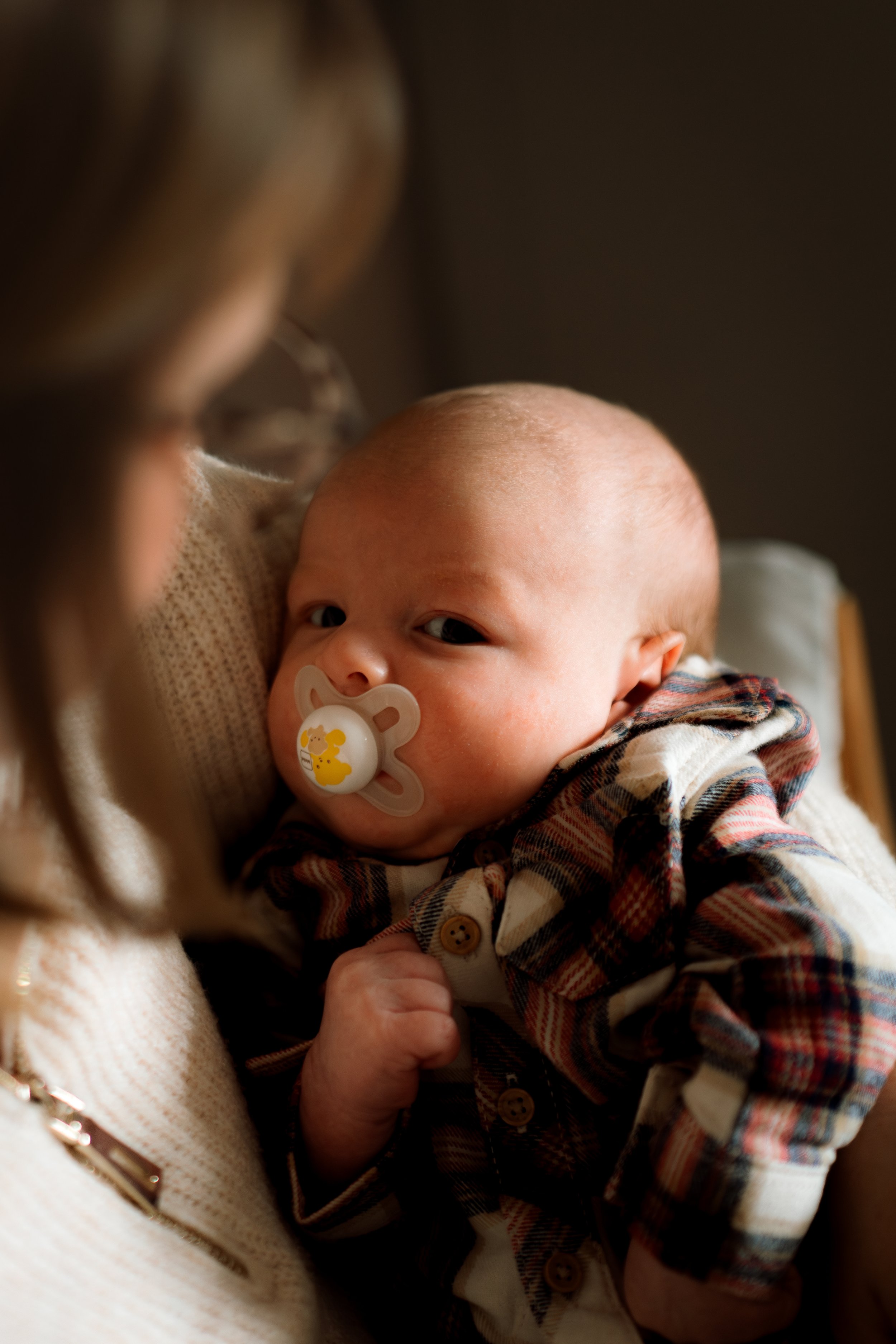 A baby with a pacifier resting on a woman's shoulder, looking at her with a sleepy expression.