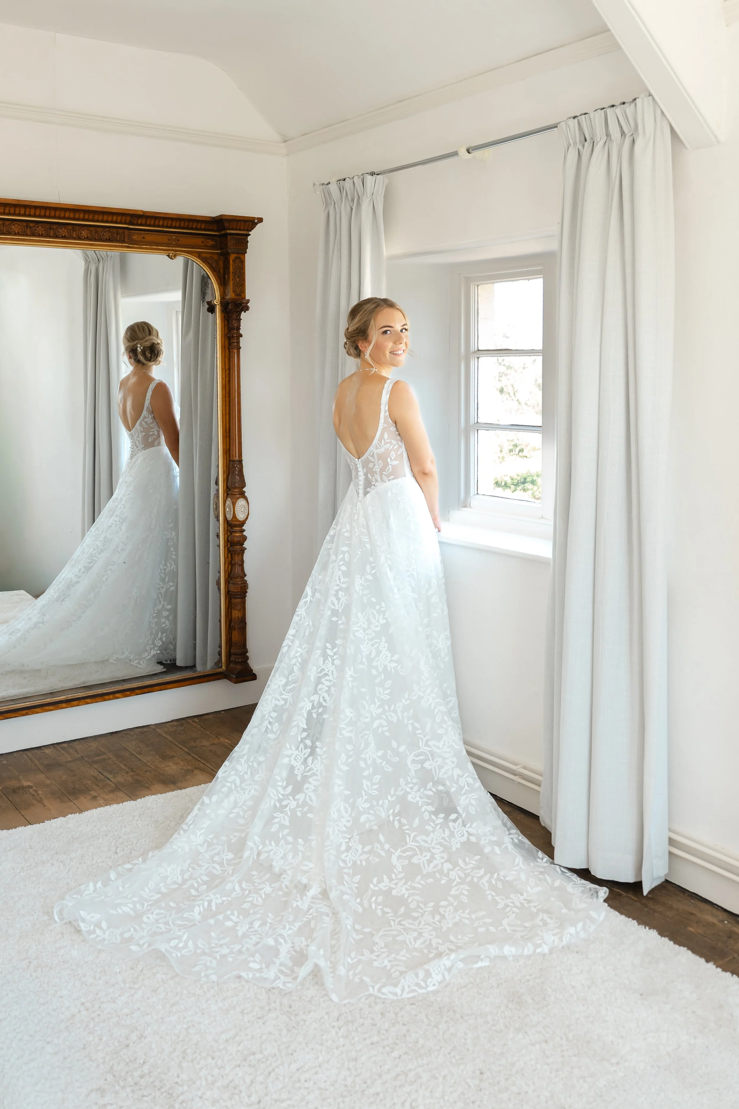 A bride in a white wedding dress with floral lace details stands near a window with white curtains, smiling at the camera, with her reflection visible in a large wooden-framed mirror behind her.