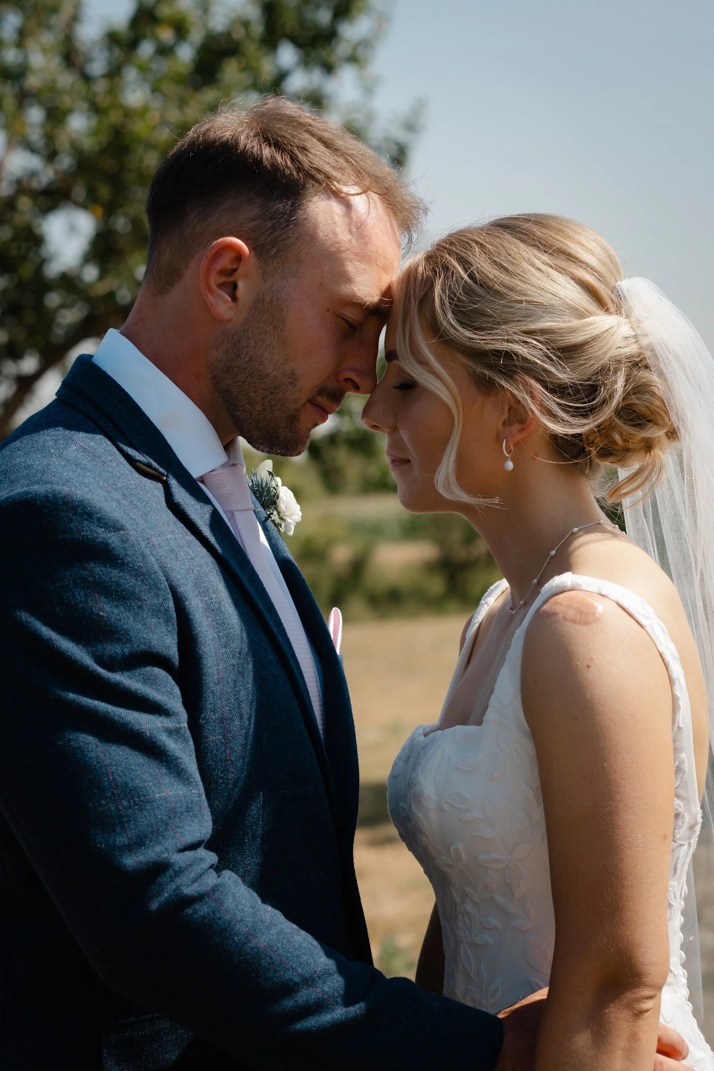 A bride and groom with foreheads touching, eyes closed outdoors on their wedding day.