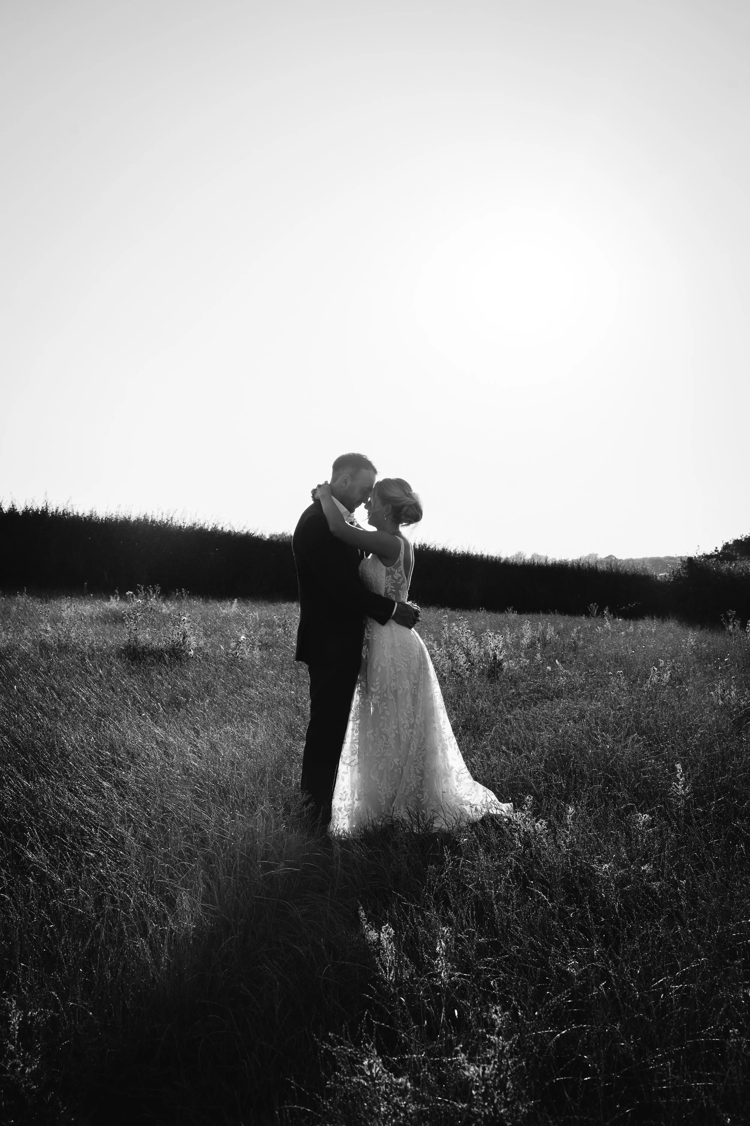 A black and white photo of a couple in wedding attire standing close together in a grassy field, with their foreheads touching and arms around each other, backlit by the sun in the distance.