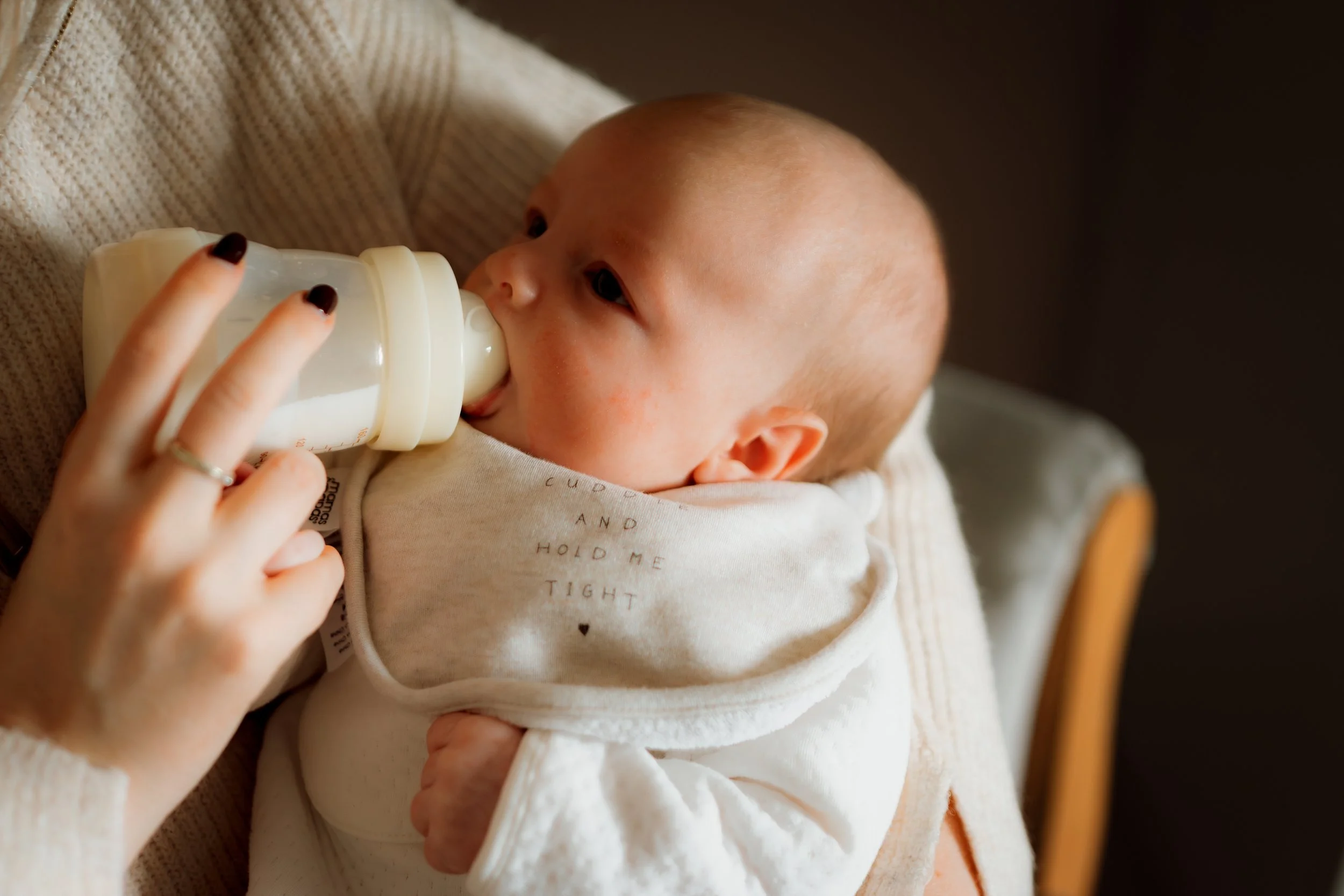 Baby lying on a cozy blanket being fed milk with a bottle held by a woman with dark nail polish.