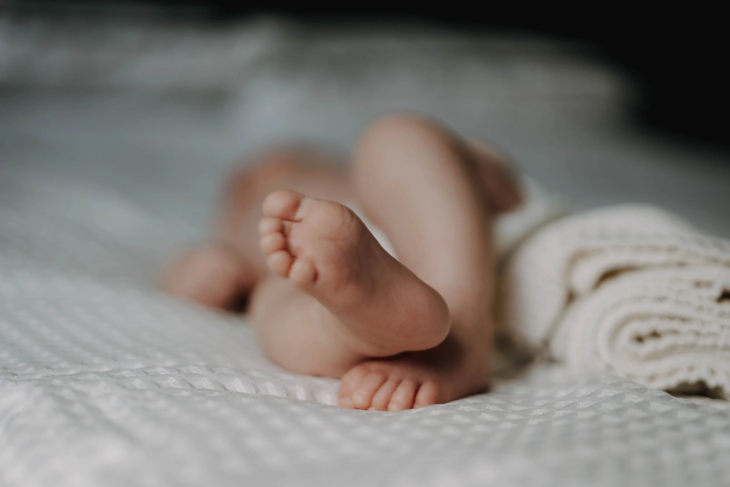 Close-up of a baby's tiny bare feet and legs, lying on a soft textured surface, with a rolled towel nearby.