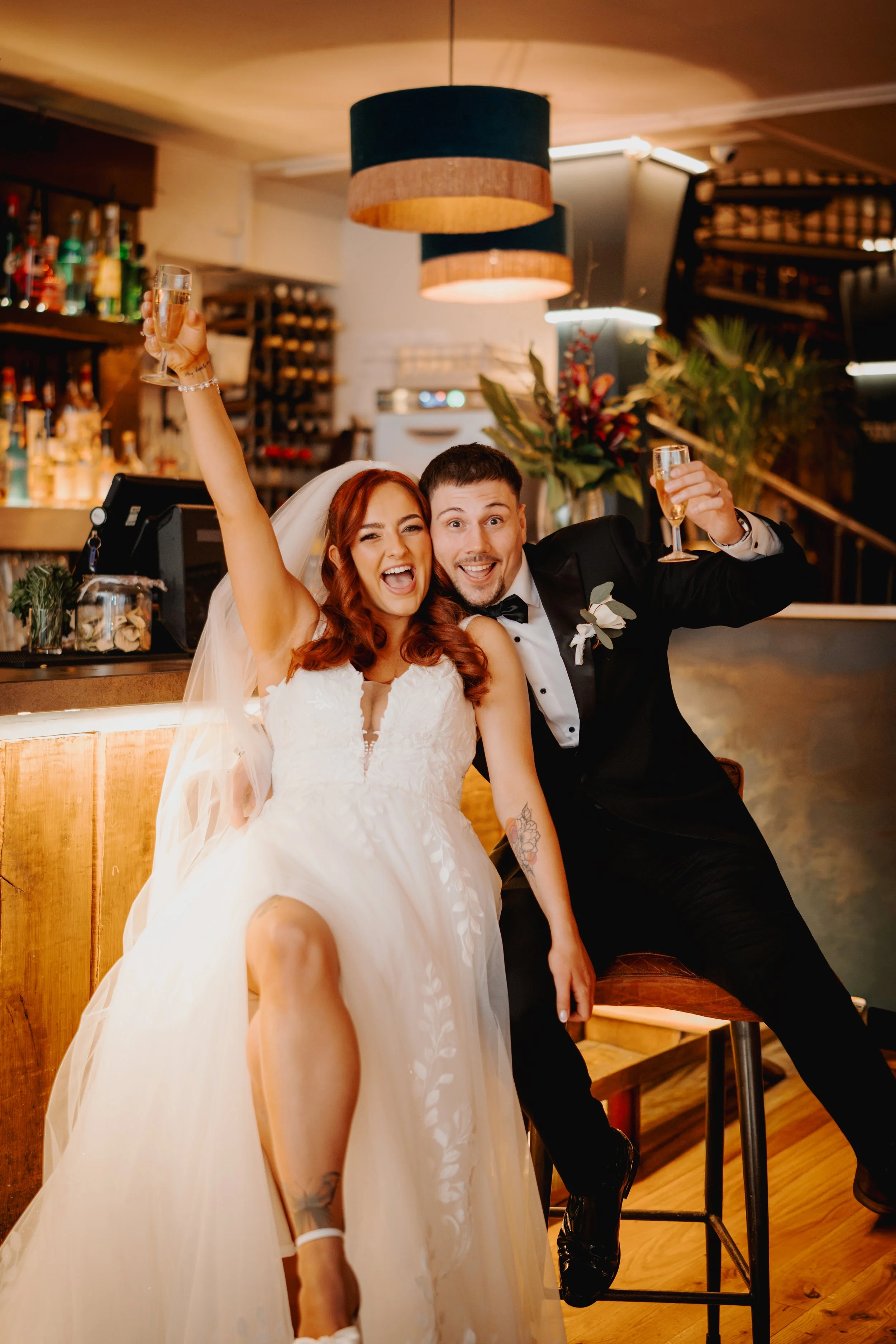 A newlywed couple celebrating with glasses of champagne in a bar setting. The bride is smiling and wearing a white wedding dress and veil, while the groom is dressed in a black tuxedo with a bow tie. They are sitting close together, with warm lightin