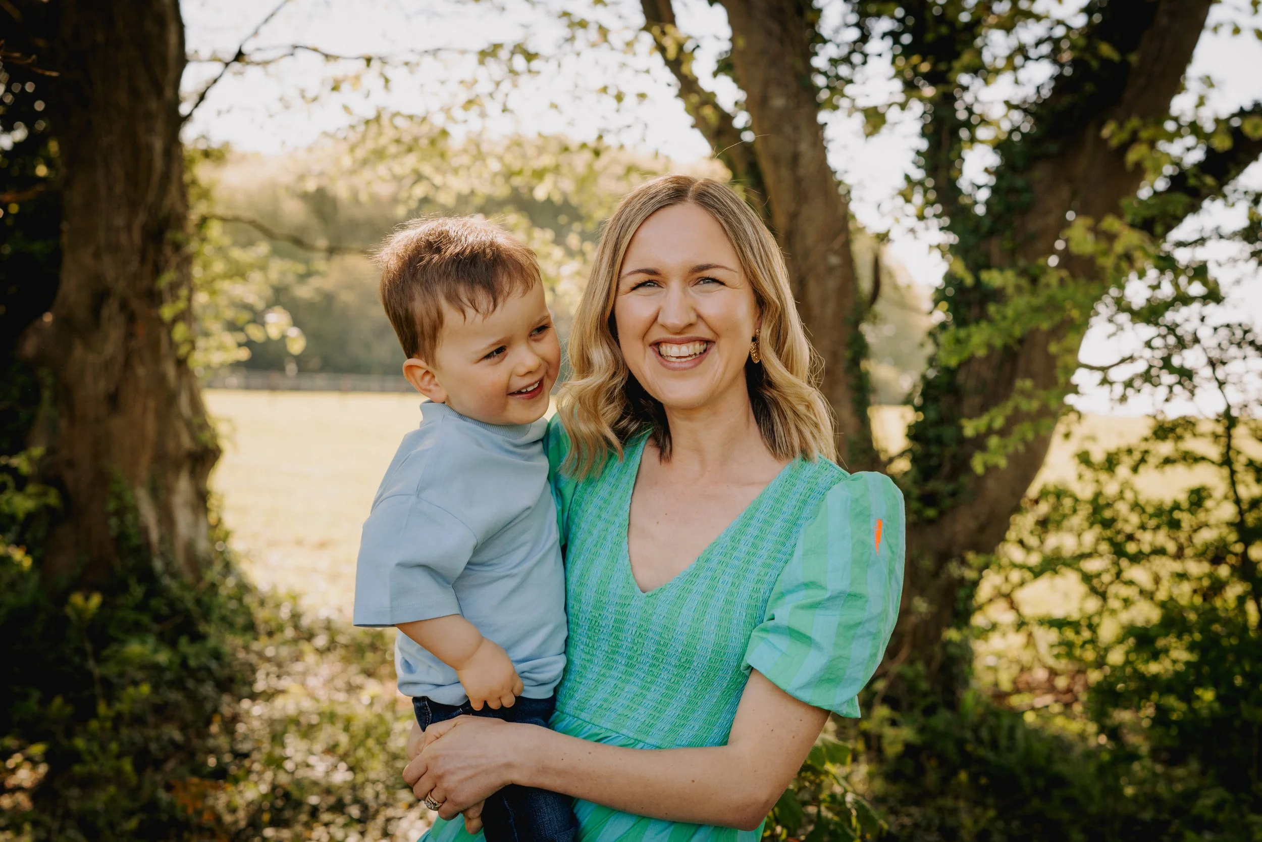 A smiling woman holding a young boy outdoors near trees in a park.