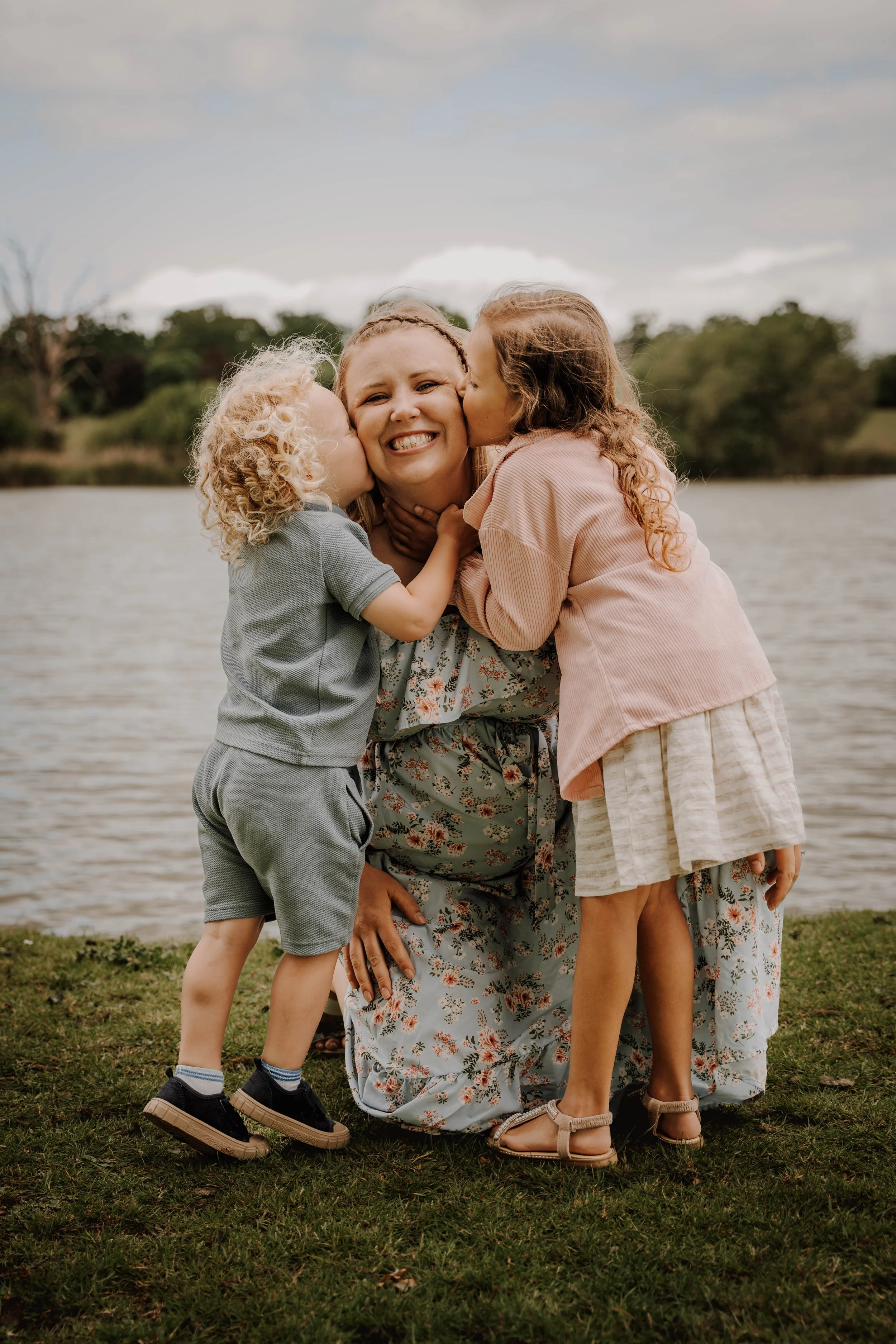 A woman sitting on the grass near a lake, smiling as two children kiss her cheeks. The background shows trees and cloudy sky.