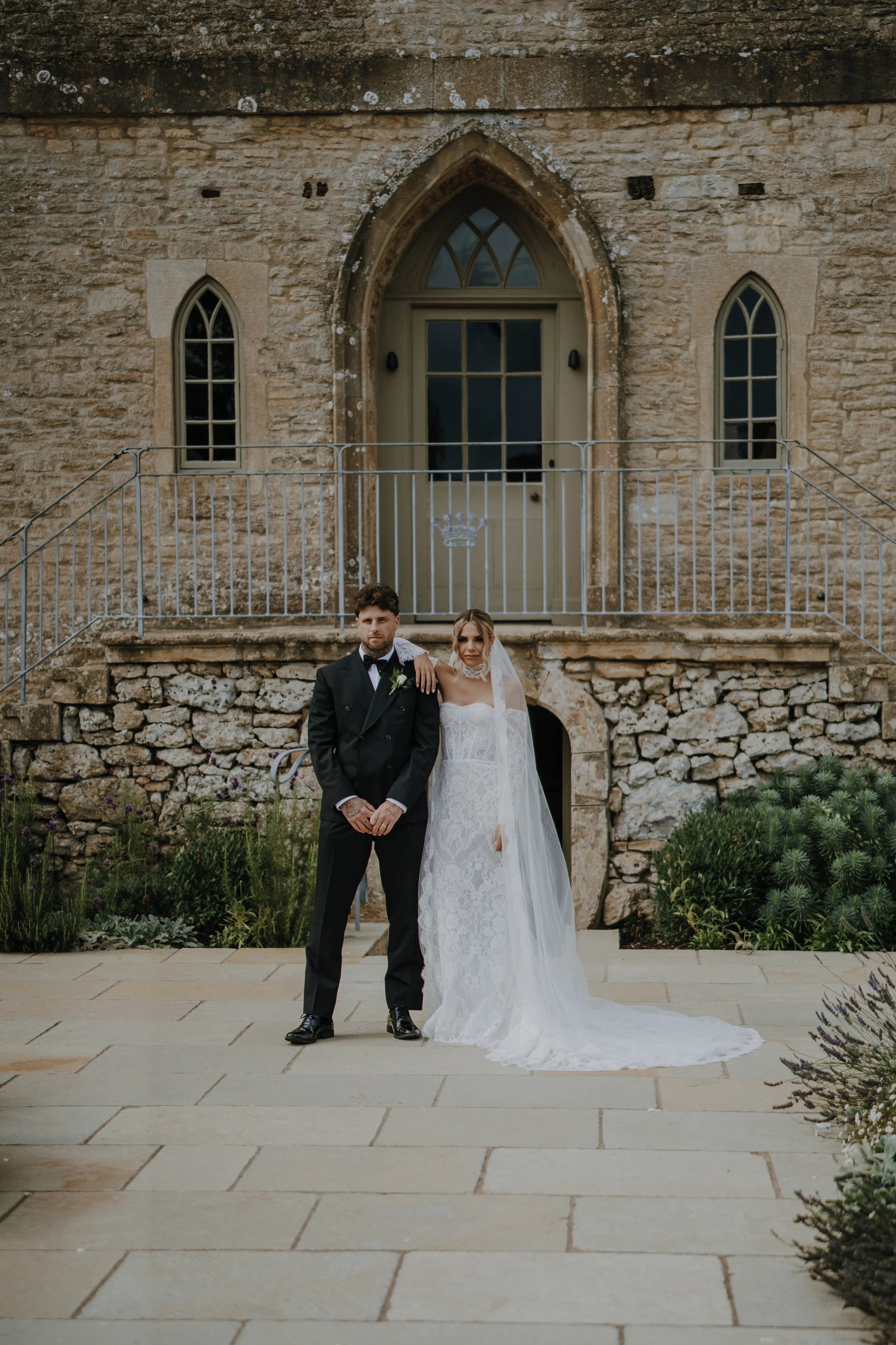 A bride and groom stand in front of an old stone building with arched windows, floral landscaping, and a metal railing, dressed in wedding attire.
