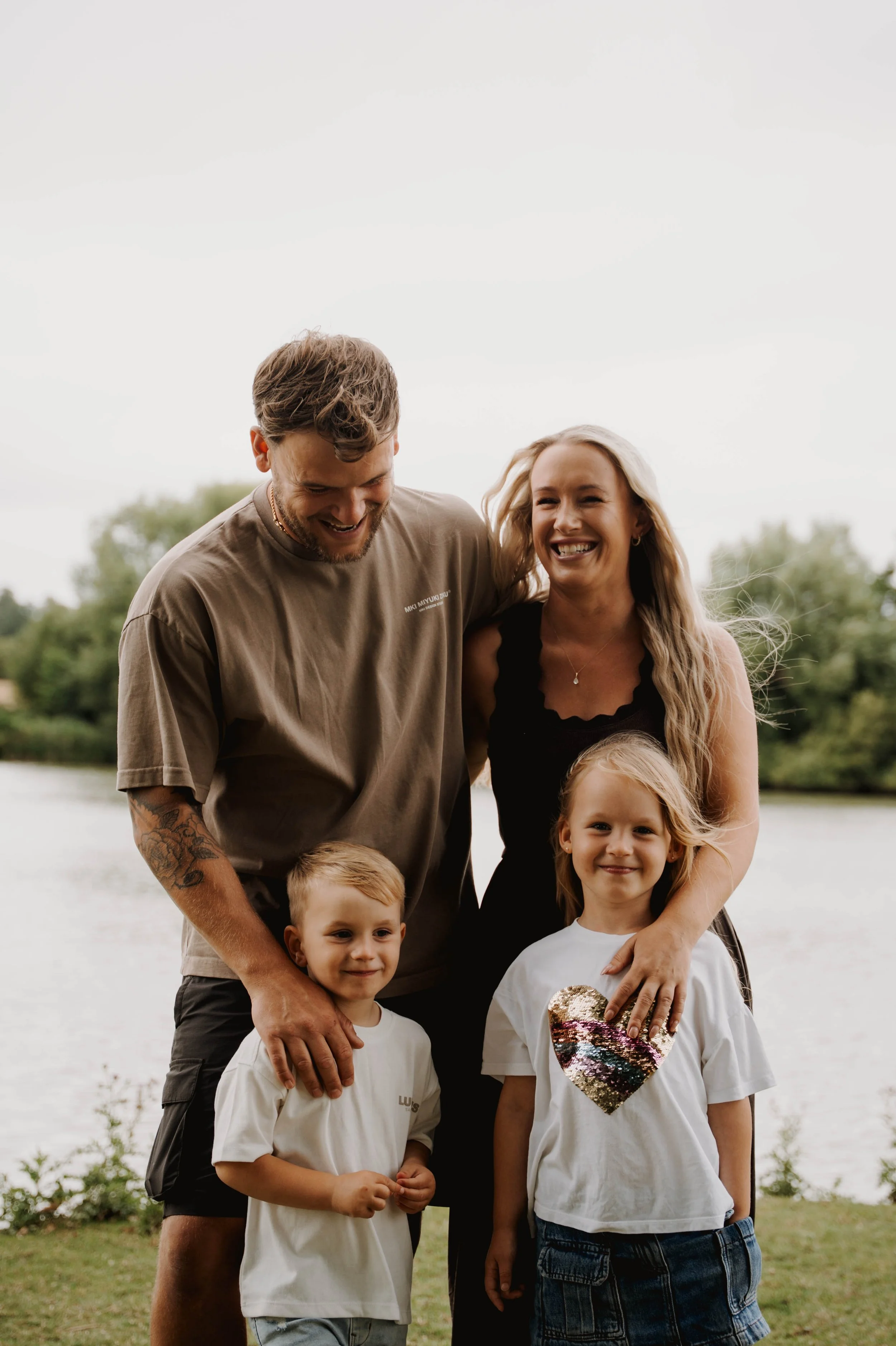 A smiling family of four stands outdoors by a river, with the father and mother hugging their two children, all enjoying a happy moment.