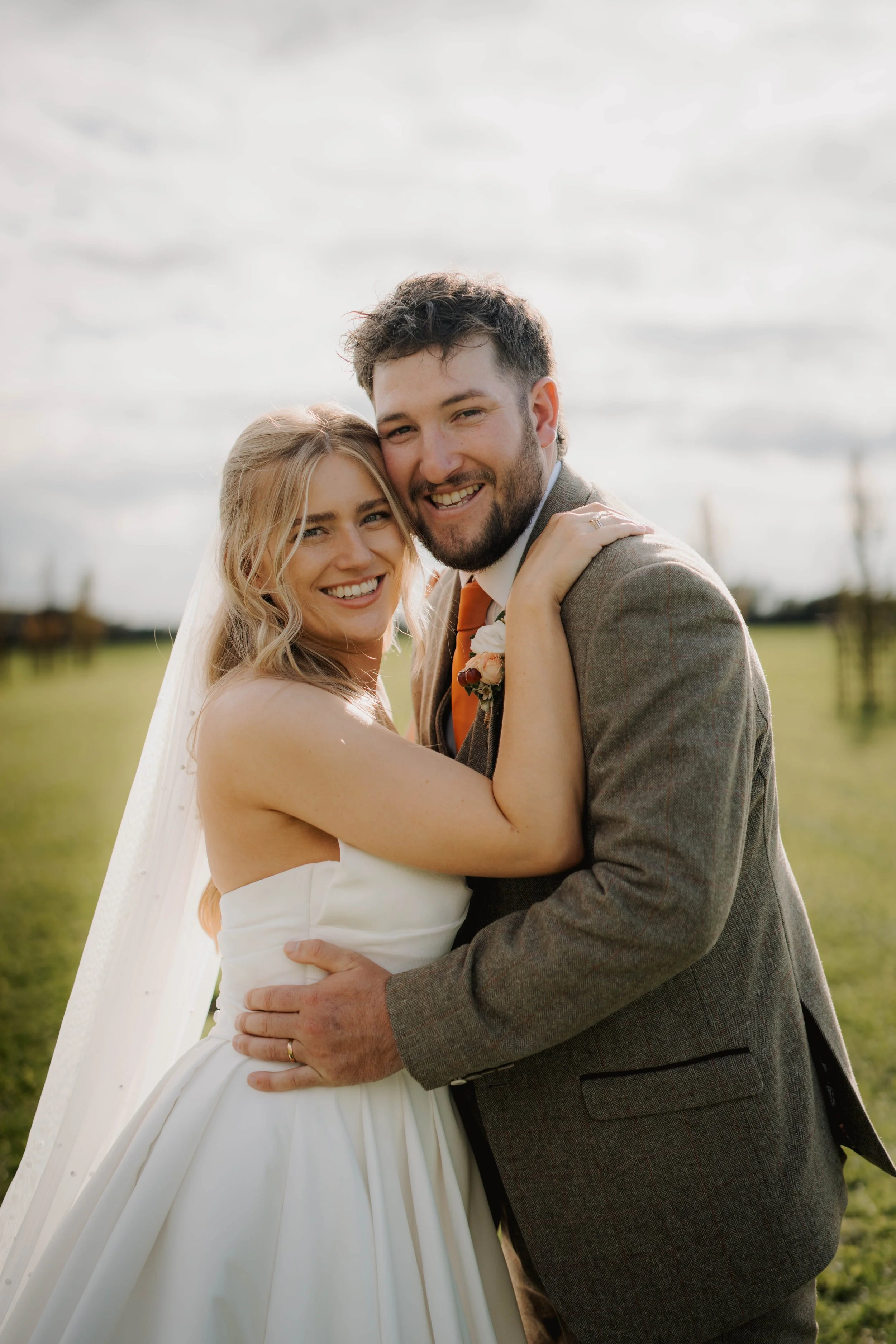 A happy couple on their wedding day outdoors, embracing each other, with a cloudy sky and grassy field in the background.