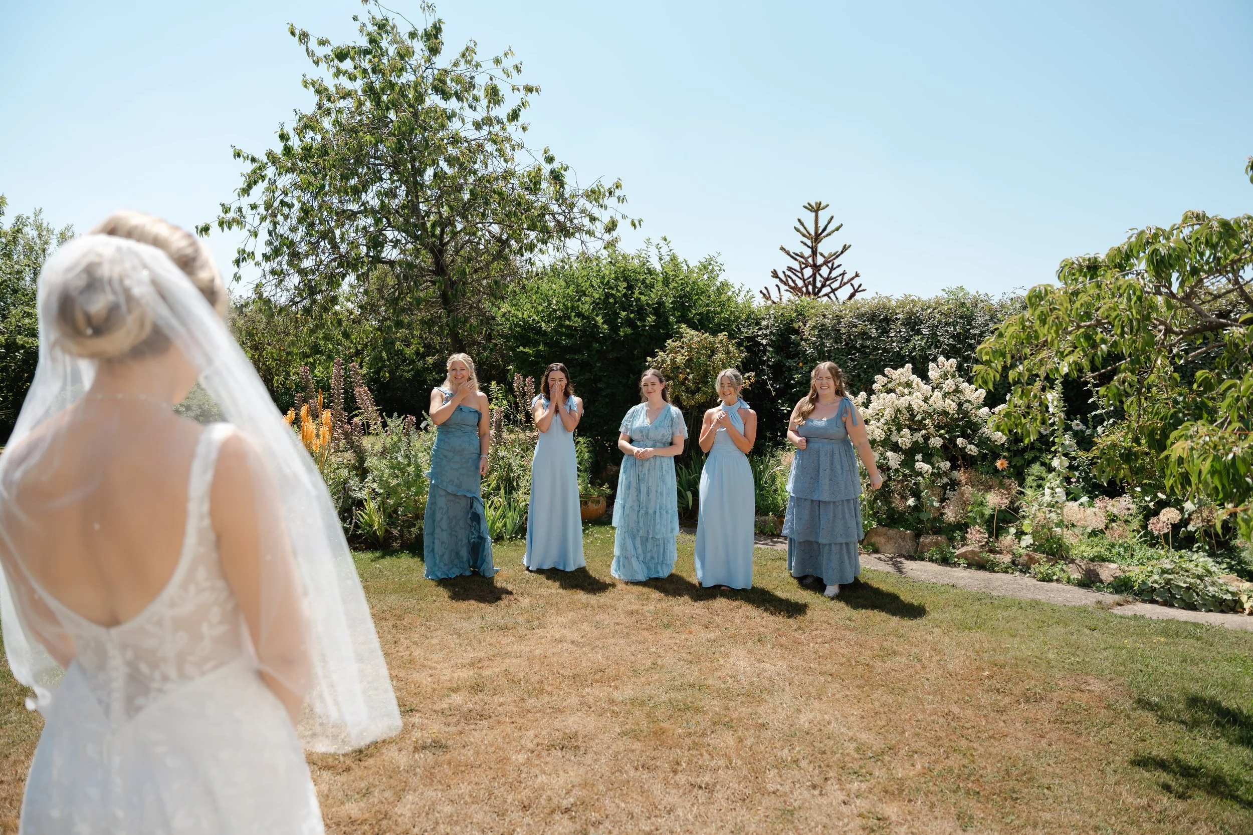A bride in a white wedding gown with lace details and veil sees five women in blue dresses, standing on a lawn with trees and flowering bushes, under a clear blue sky.