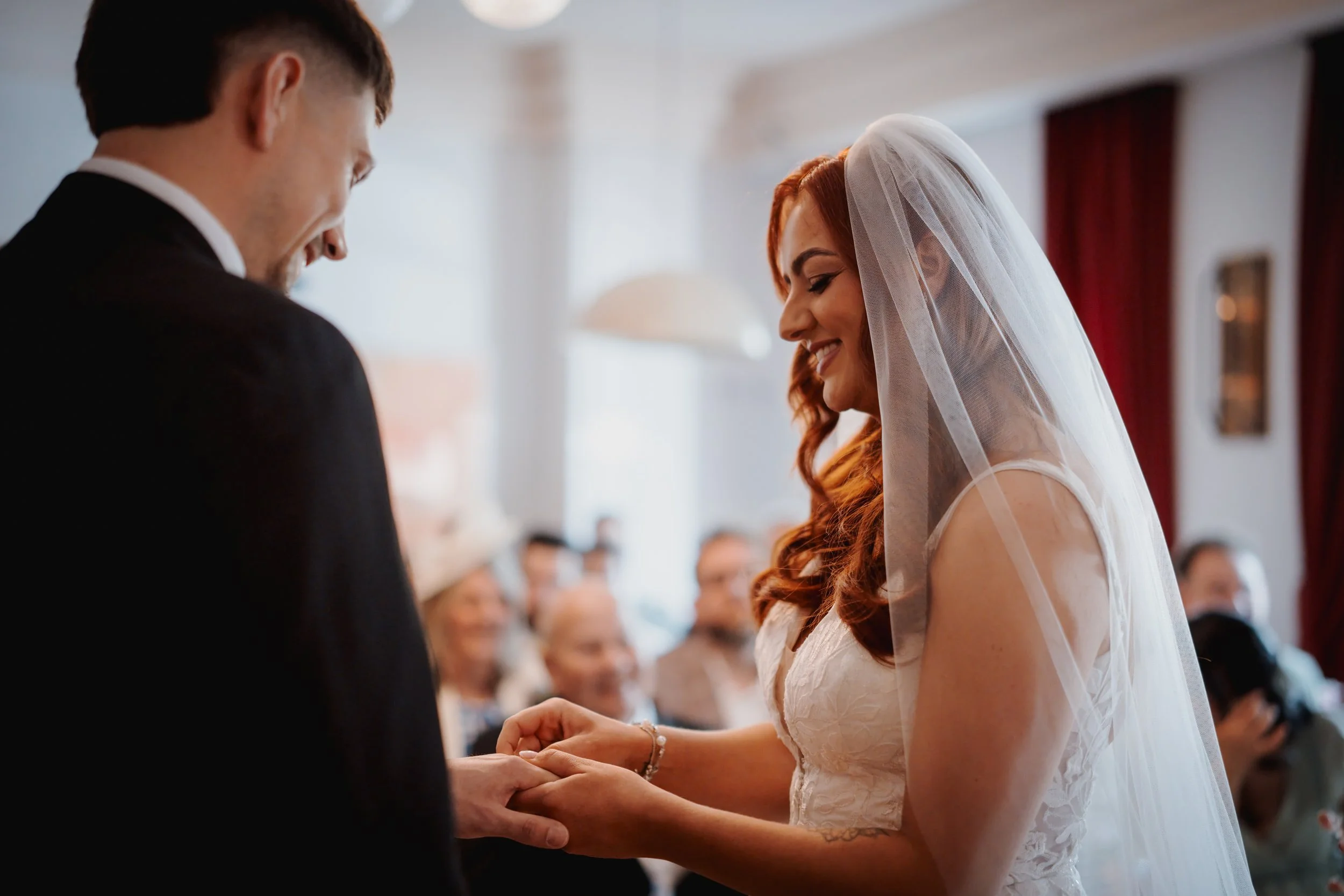 A bride and groom exchanging wedding vows and holding hands during their wedding ceremony. The bride wears a white lace dress and a veil, and the groom is dressed in a black suit. Guests are visible in the background.