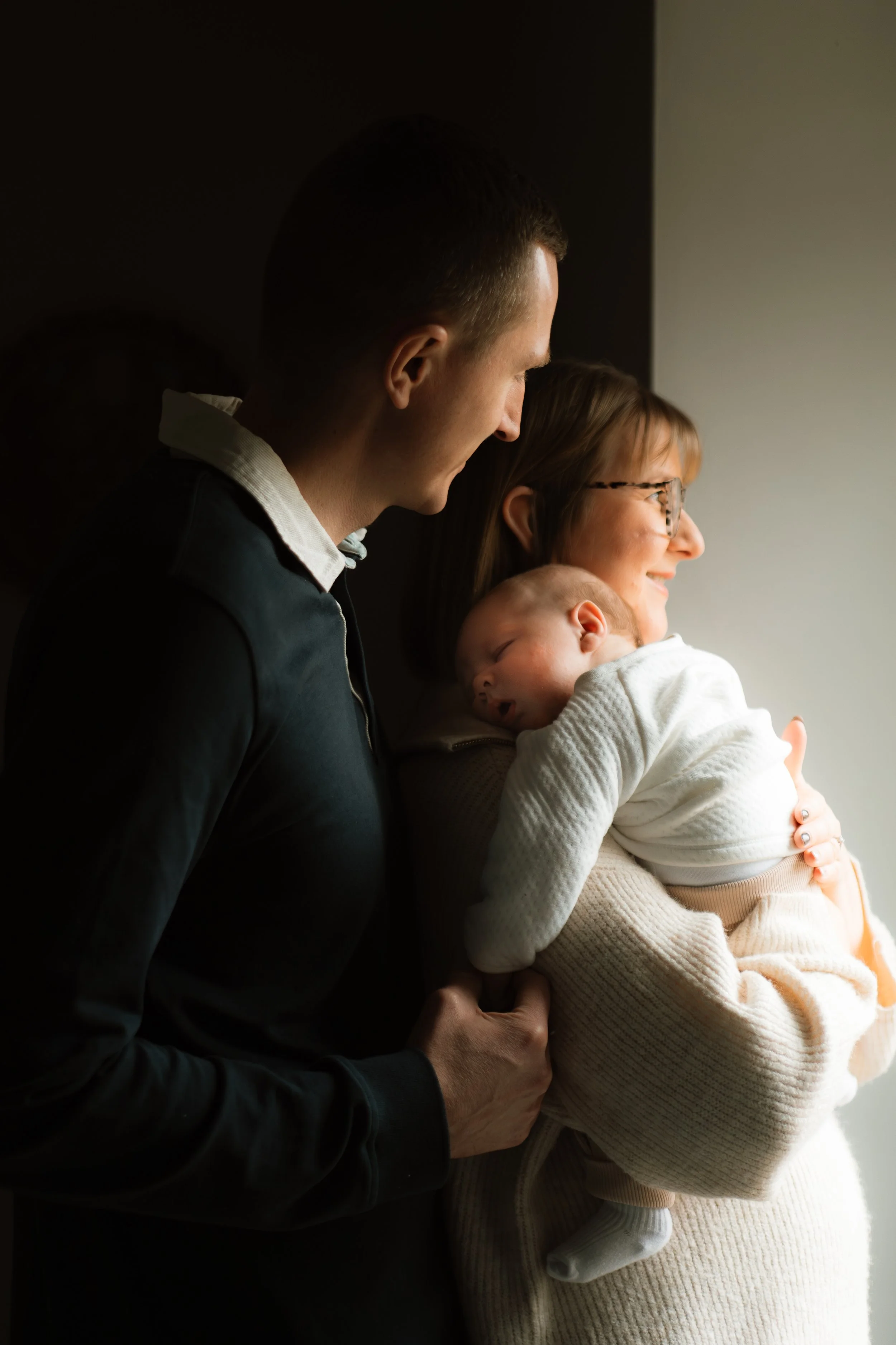 A family of three, a man, a woman, and a sleeping baby, looking out a window with soft natural light illuminating their faces.