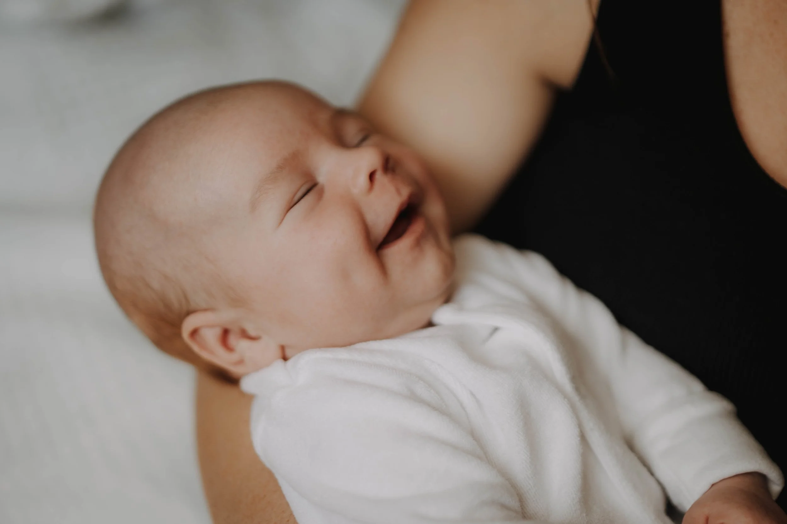 Smiling baby with closed eyes lying on a woman's chest, both appearing happy and relaxed.
