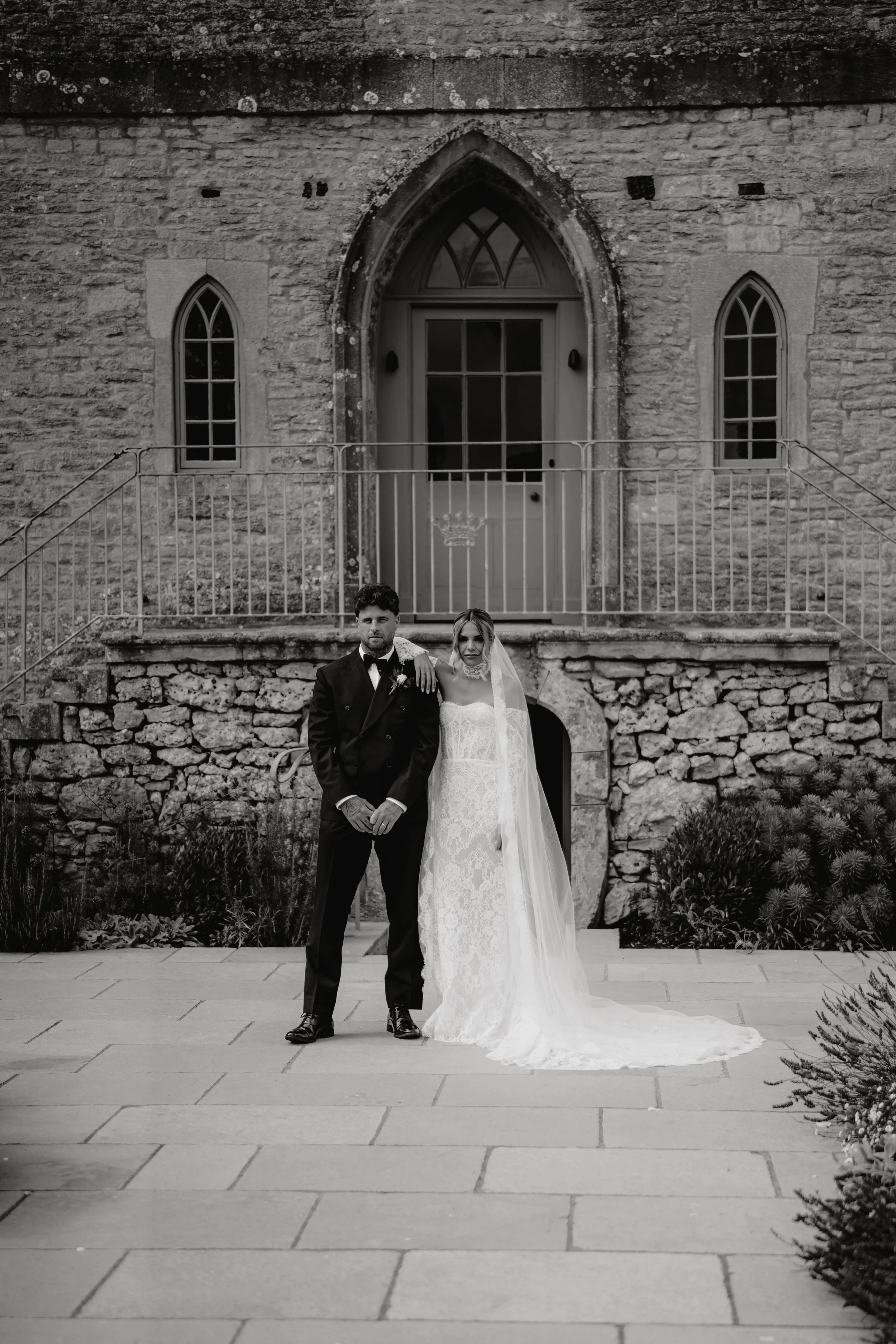 Black-and-white photo of a bride and groom standing in front of an old stone building with arched windows and a wooden door, the groom in a tuxedo and the bride in a lace wedding gown with a veil.