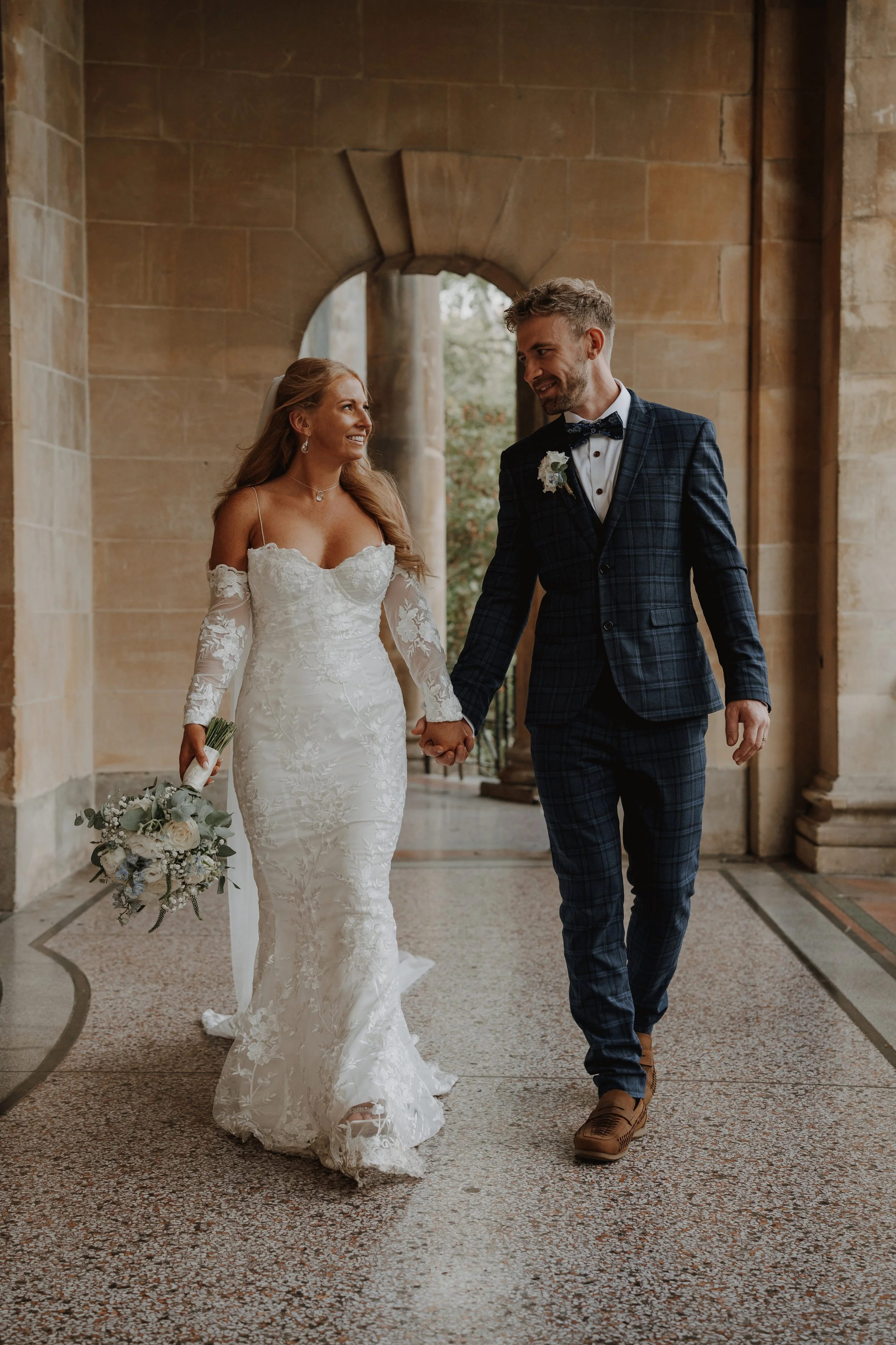 Bride and groom holding hands, walking through a historic stone building, smiling at each other, woman in a lace wedding dress with floral details, man in a dark plaid suit with a bow tie, wedding bouquet in woman's hand.